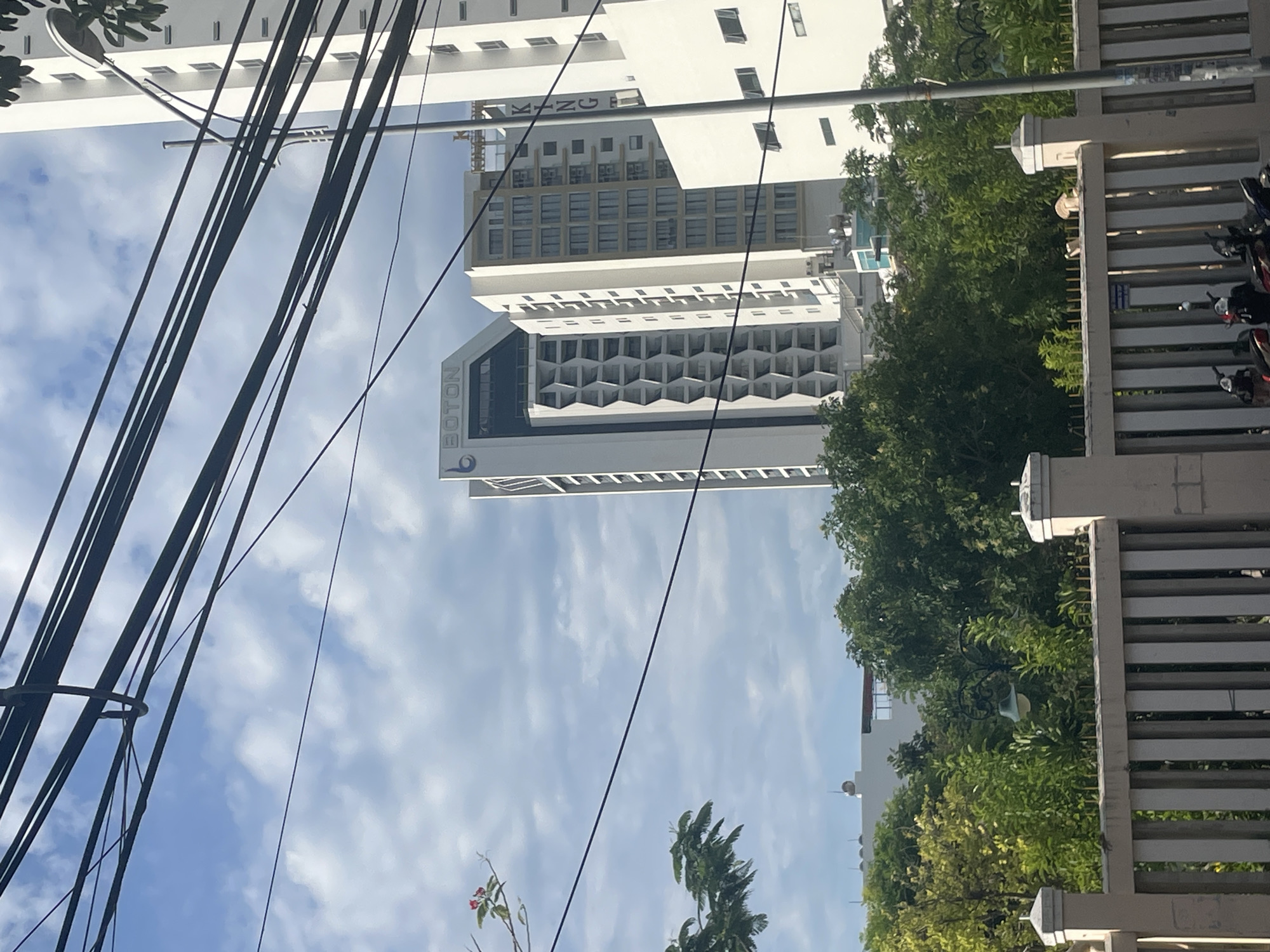 In Nha Trang, Vietnam, a modern cityscape unfolds under a cloudy sky, bisected by a network of dark electrical wires. Dominating the frame are several tall, contemporary buildings. One prominent structure, labeled "BOTON," features a distinctive facade with hexagonal patterns. Adjacent to it stands another white building with a grid of windows. To the right, a taller white building is visible, with the word "KING" vertically displayed on its side. In the foreground, a beige fence with vertical slats separates a street scene from the buildings. Several motorcycles are parked behind the fence, suggesting activity in the area. Lush green trees and foliage are interspersed between the buildings and in the foreground, softening the urban landscape. The lighting indicates it is daytime.