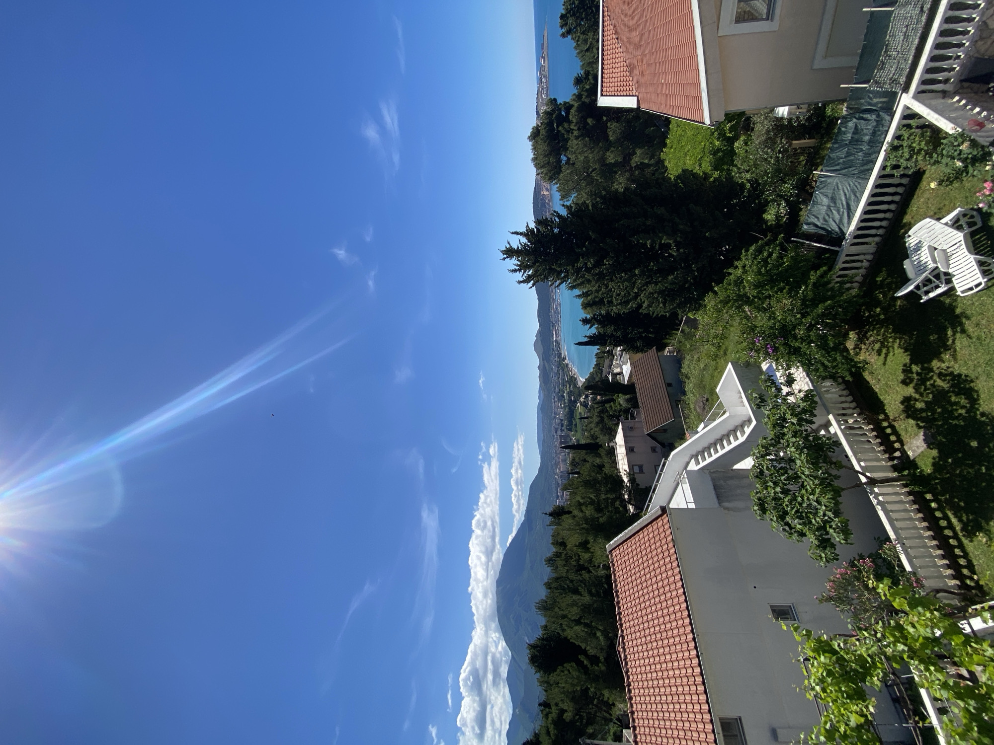 A vibrant, sunlit landscape is captured from an elevated perspective, showcasing a coastal town backed by mountains and extending into the sea, consistent with the location of Šušanj, Montenegro.
The upper portion of the image is dominated by a clear, brilliant blue sky with faint, wispy white clouds. The sun shines brightly from the top-left corner, creating a noticeable lens flare and sunburst effect. A tiny dark object, possibly a bird, is visible against the sky near the sun.
In the mid-distance, a long stretch of turquoise sea meets a bustling coastline, dotted with buildings of a town or city. Behind this, imposing mountains rise, their peaks sometimes veiled by white cumulus clouds. Lush green forests cover the slopes of the mountains and the hills leading down to the coast.
The immediate foreground features several residential buildings with distinctive red-tiled roofs and light-colored facades. White balustraded staircases descend through verdant gardens. A neatly maintained grassy lawn is visible, complete with white outdoor lounge chairs. Various green trees, bushes, and grapevines are scattered throughout the gardens, casting dappled shadows. A dark green tarp covers an area near one of the buildings.
The bright sunlight suggests either morning or afternoon. No specific activity or event is taking place; it is a static scenic view. No visible text is present in the image.