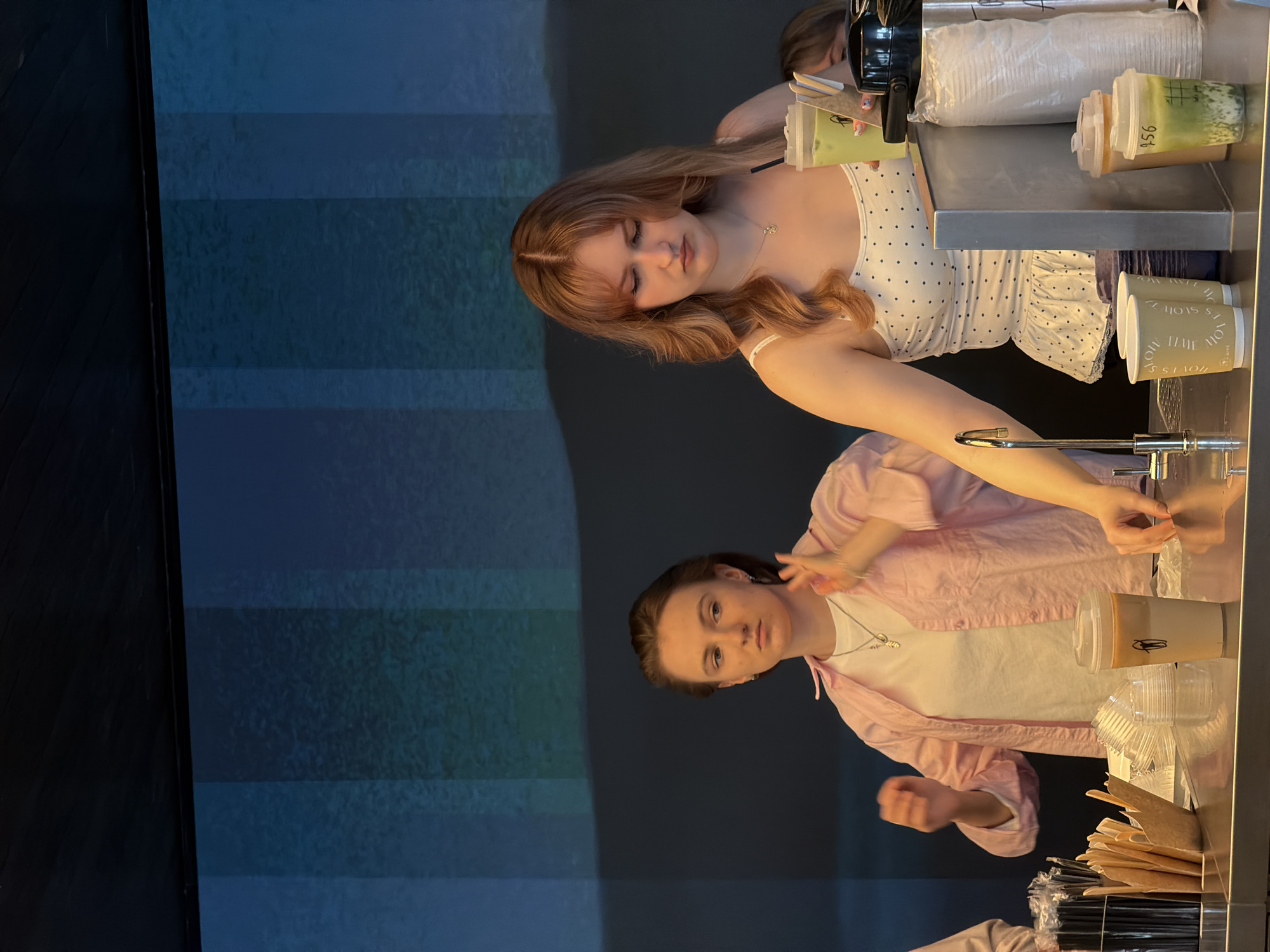 Two people are at a drink station. The first person, with strawberry blonde hair and wearing a white top with black polka dots, is making a green tea beverage. The second person, with dark hair and wearing a pink shirt over a white shirt, is preparing another beverage. The environment appears to be the inside of a cafe. There are cups, straws, and other beverage-making supplies visible at the drink station. The location in Moscow, Russia, is unknown.
