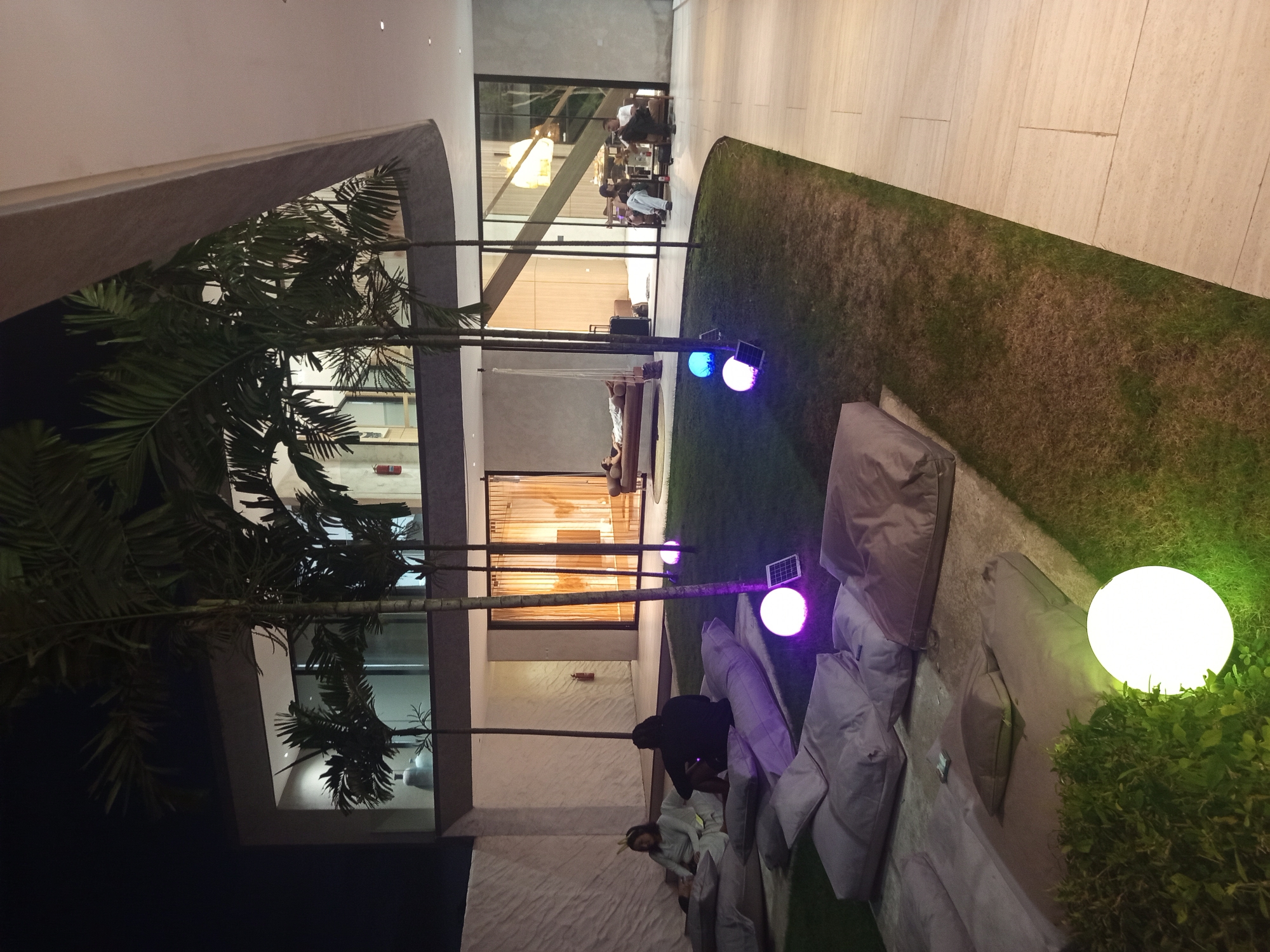The image shows an outdoor area in Kuta Selatan, Indonesia, at night. Several beanbag chairs in light gray are scattered on a grassy area. A couple of people are near the beanbag chairs. There are several spherical lamps, some with colored light and some white. A modern building with large windows and trees is visible in the background.