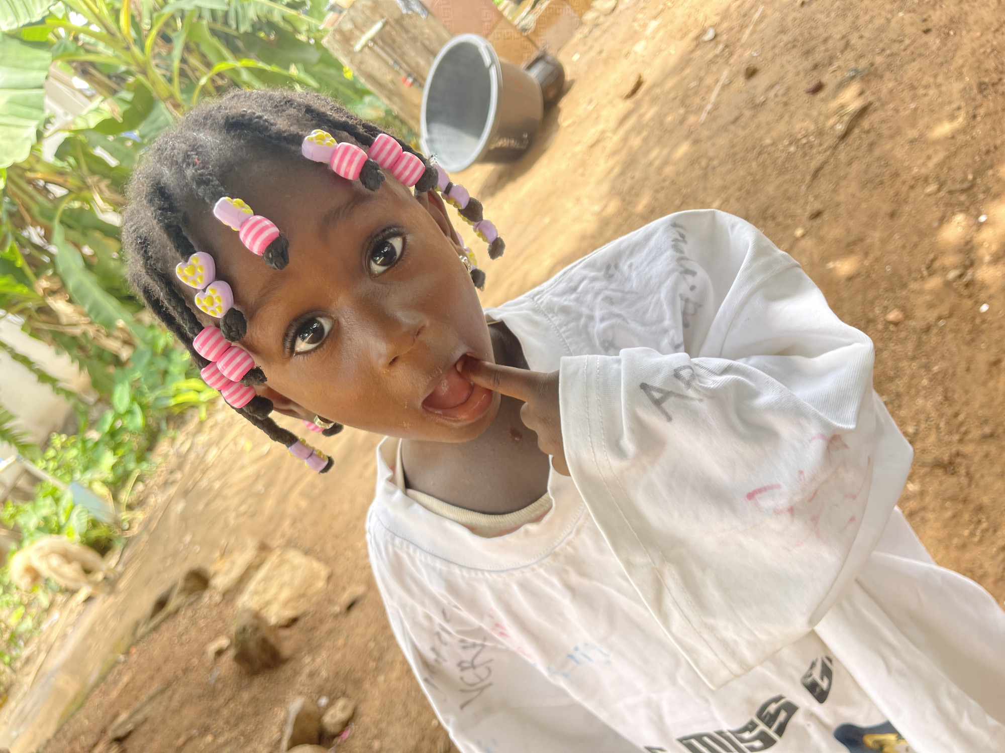 The image features a young Black child, likely female, captured from a slightly low and tilted perspective, looking directly into the camera. She has dark, braided hair adorned with numerous colorful beads, including pink and white striped cylindrical beads and light purple heart-shaped beads with yellow centers. Her eyes are wide and dark, and her mouth is slightly open with her right index finger placed inside.
She is wearing an oversized, loose-fitting white t-shirt, which appears to have some faded writing or marks on it. Visible text includes "AR" on her left sleeve and "MISS" faintly near the bottom right of the shirt.
The setting is outdoors, characterized by dry, reddish-brown earthy ground with scattered small stones and debris. Lush green foliage, possibly large-leaf plants like bananas, is visible in the background on the left, indicating a vegetated area. In the upper right background, a silver-colored bucket and another dark object rest on the ground. The bright lighting suggests it is daytime. The scene conveys a candid moment, capturing the child's curious and perhaps playful expression in an informal outdoor environment in Akwanga, Nigeria.