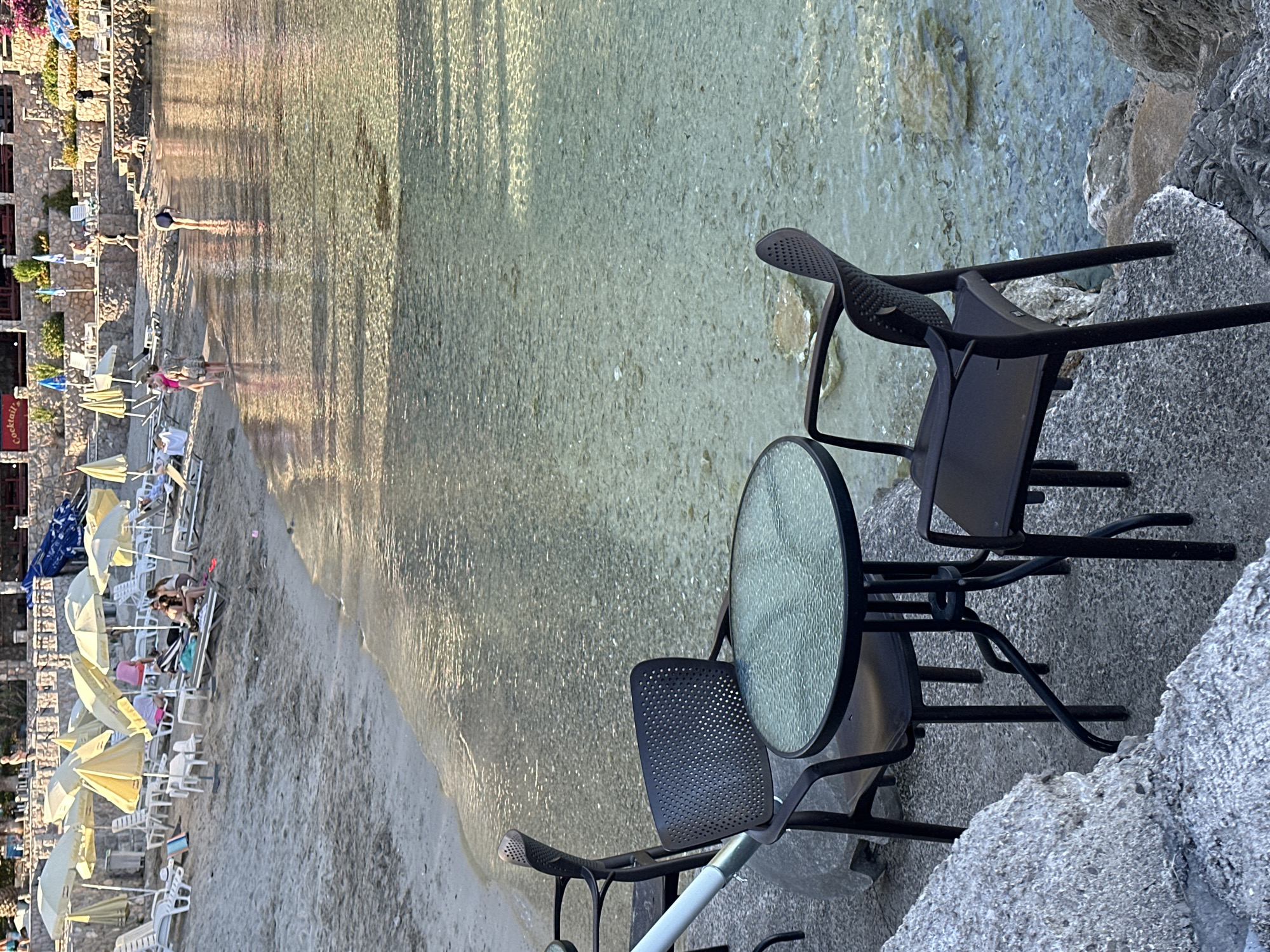 The image displays a coastal scene in Seoca, Montenegro, under warm, low-angle sunlight. In the immediate foreground, a round table with a textured glass top is positioned between two dark-colored plastic chairs, each with a perforated backrest. This furniture rests on a grey, pebbled concrete surface situated at the very edge of calm, clear, shallow water. An irregular grey rock structure borders the concrete on the left.
The water, extending from the foreground, reveals a light-colored, rocky bottom and reflects sunlight, creating shimmering patterns. To the left of the water, a light-tan sandy beach is occupied by numerous open yellow and white beach umbrellas, beneath which multiple white plastic lounge chairs are arranged.
Several individuals are present within the scene. In the shallow water, one person wearing a dark swimsuit is bent over, facing away from the viewer. On the sandy edge, two individuals stand facing each other: one in a pink swimsuit and another in a patterned dress. Approximately 5-8 additional people are indistinctly visible relaxing on lounge chairs under the beach umbrellas.
In the background, a multi-tiered stone structure with green foliage ascends from the beach. A building on the uppermost tier features a red sign prominently displaying the word "Cocktails." Additional people are discernible walking or standing on these higher terraces.