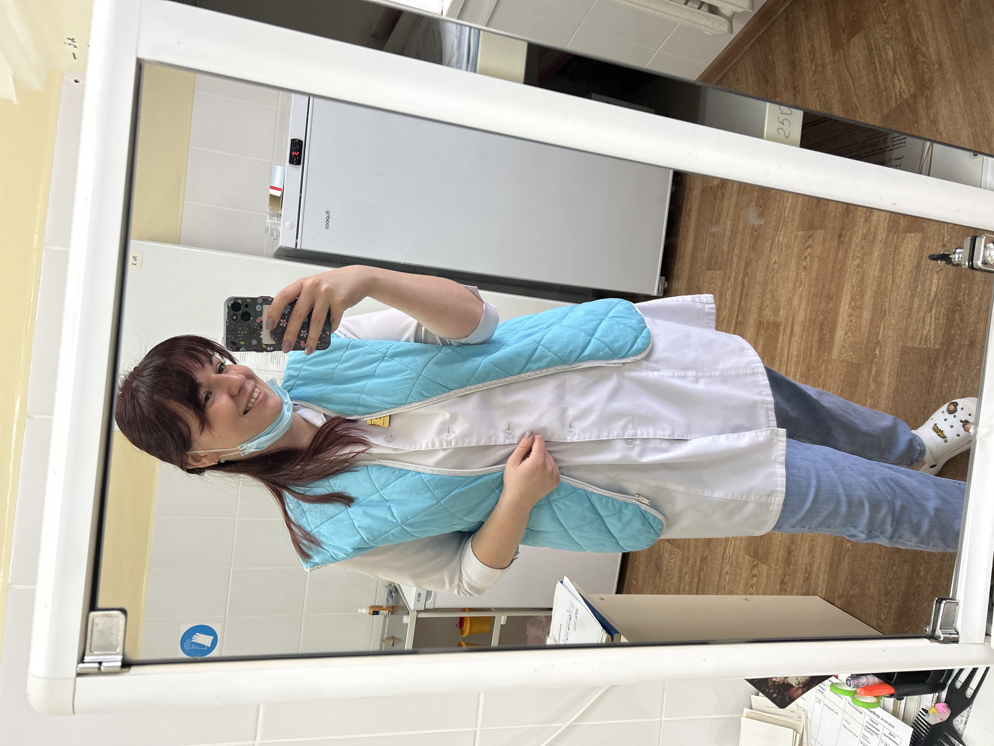 A woman wearing a medical coat and a light blue quilted wrap is taking a selfie in a mirror. She is wearing a blue disposable face mask. The reflection shows a white refrigerator, wooden flooring, and a tiled wall with a sticker of a blue glove. A document titled "Утверждаю Щеглов Впр 28.04.2025" is visible in the lower right corner of the mirror. The mirror's location is a medical facility in the city of Фоки, Russia.