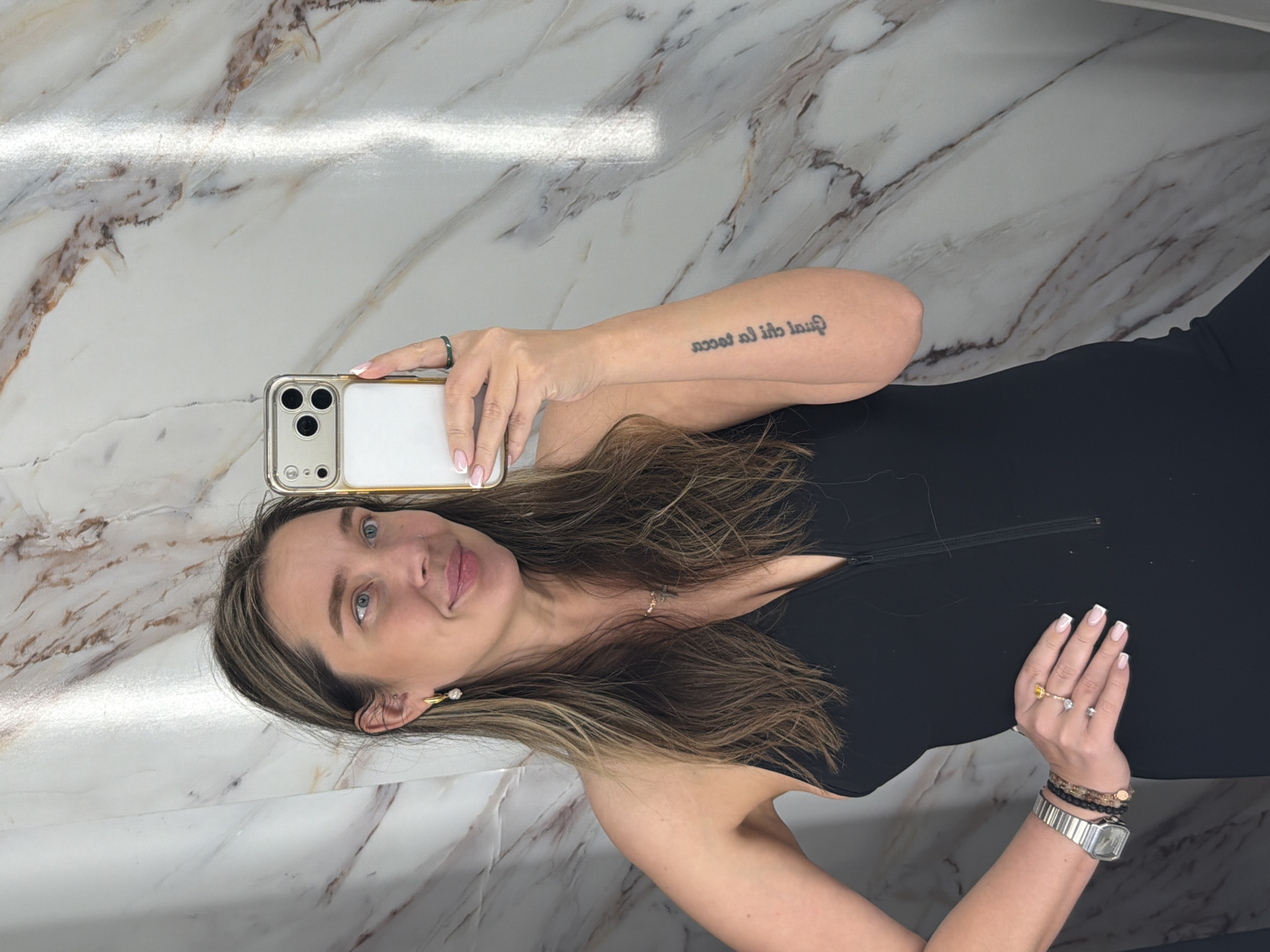 A woman with brown hair and blue eyes is taking a selfie. She is wearing a black dress, earrings, and a necklace. She has a tattoo on her right arm. The background is a marble wall. This is not in Moscow, Russia.