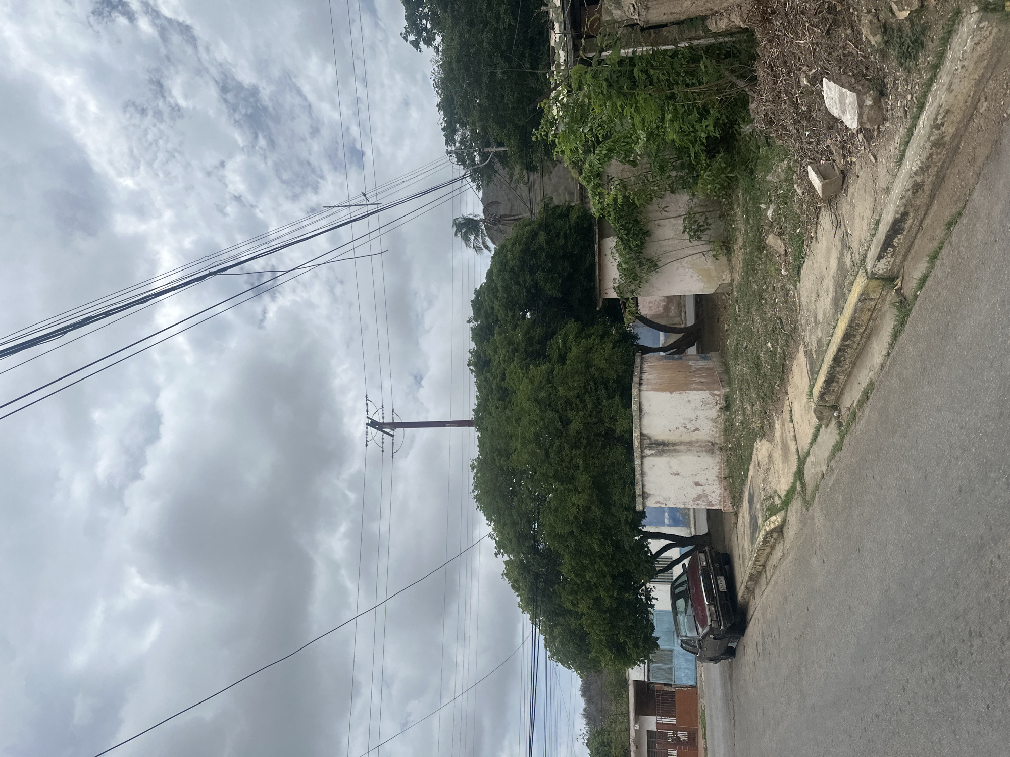 The image depicts a street scene in El Maco, Venezuela, under an overcast sky. A dark-colored sedan is parked on the right side of the street, partially on the sidewalk. In the background, several low-rise structures are visible, along with lush green trees and vegetation. Utility poles and a network of power lines crisscross the upper portion of the frame. The overall impression is of a somewhat weathered, urban environment.