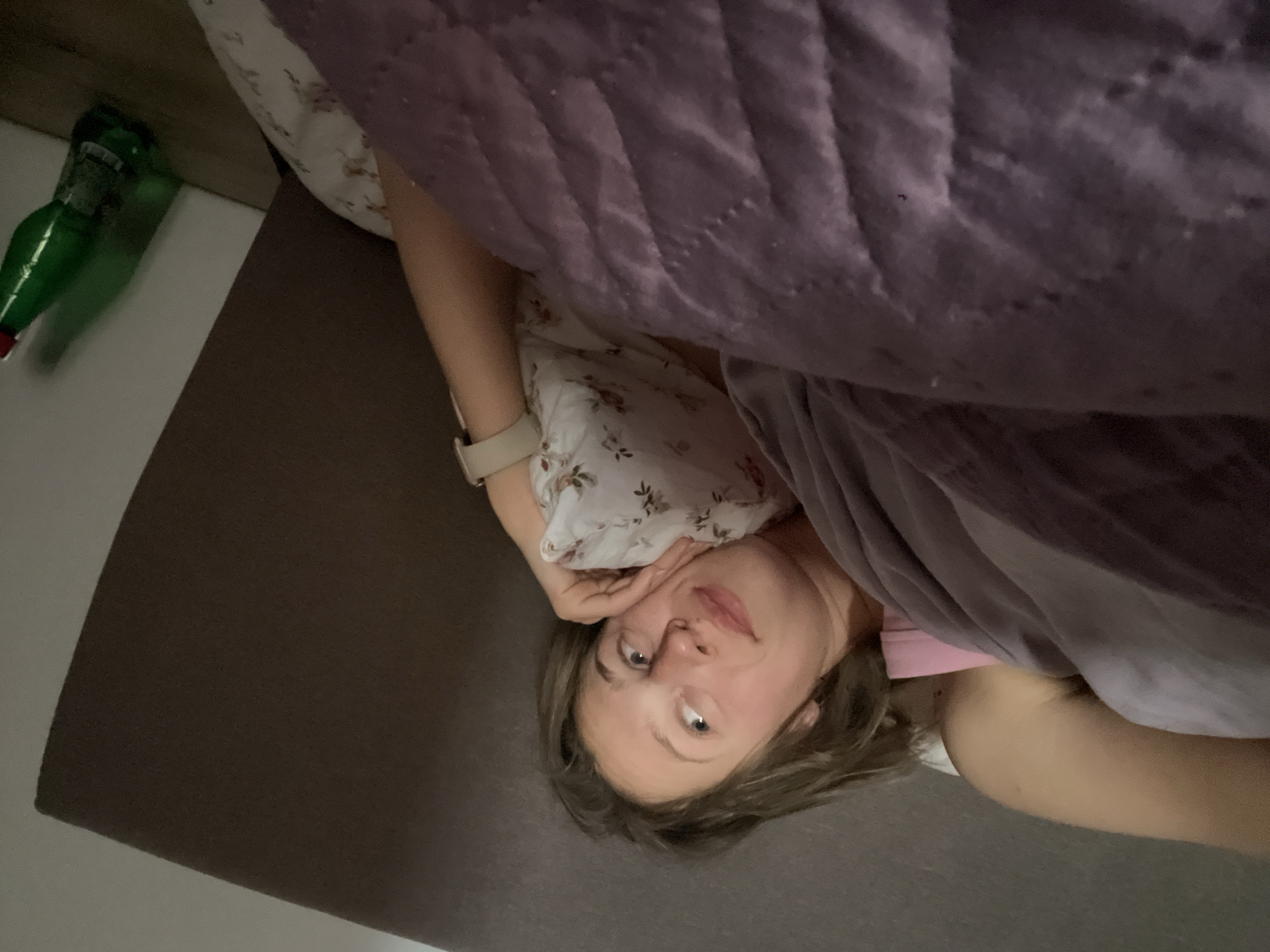 A person is lying on a bed, partially covered by a purple quilt. They have fair skin, light-colored hair, and are wearing a light-colored floral top. Their head is tilted to the side, and their hand is near their face. A green bottle is visible on a surface to the left. The scene is indoors. No location is indicated to confirm whether this image was shot in Bečići, Montenegro.