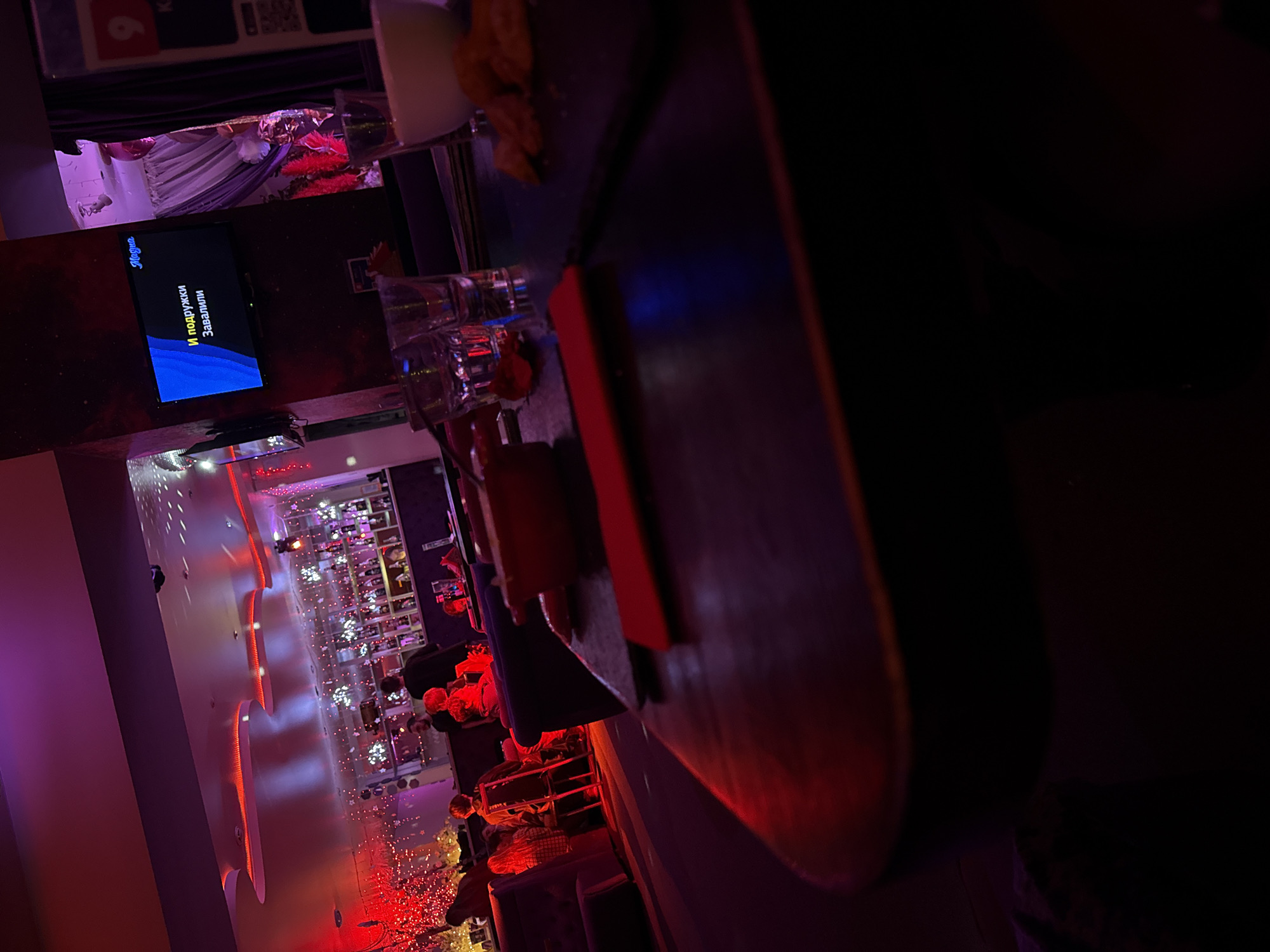 The image depicts the interior of a karaoke bar in Votkinsk, Russia. The perspective is from behind a bar counter, which dominates the right side of the frame. The counter is dark and partially obscures the view.
On the counter, there are several glasses and a container of what appears to be snacks. A screen on the wall displays karaoke lyrics in Russian, including the word "Музыка".
The bar extends into the background, with tables occupied by people. The space is decorated with lights and festive decorations.