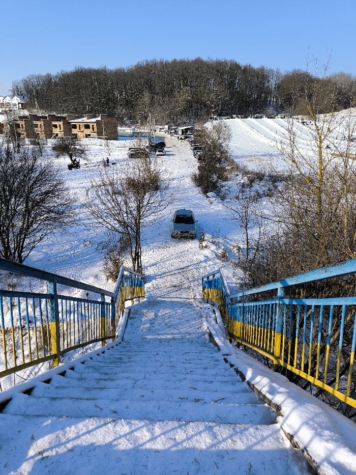 The image displays a snow-covered winter landscape in Prybuzke, Ukraine, under a clear blue sky. In the foreground, a set of outdoor steps, coated with snow, descends into the scene. The metal handrails on either side are painted in alternating blue and yellow stripes, indicative of the Ukrainian flag colors. Below the stairs, a light-colored van is parked on a snow-covered path, facing away from the viewer. To the left, several multi-story brick buildings, appearing to be under construction, are visible. An all-terrain vehicle (ATV) and a few individuals are present on the snow in front of these buildings. Further into the background, a recreational snow tubing or skiing slope with multiple parallel tracks is visible on the right, populated by several individuals. Numerous parked vehicles line a snowy road traversing the scene towards the base of the slope. A dense forest of bare, deciduous trees forms the distant background.