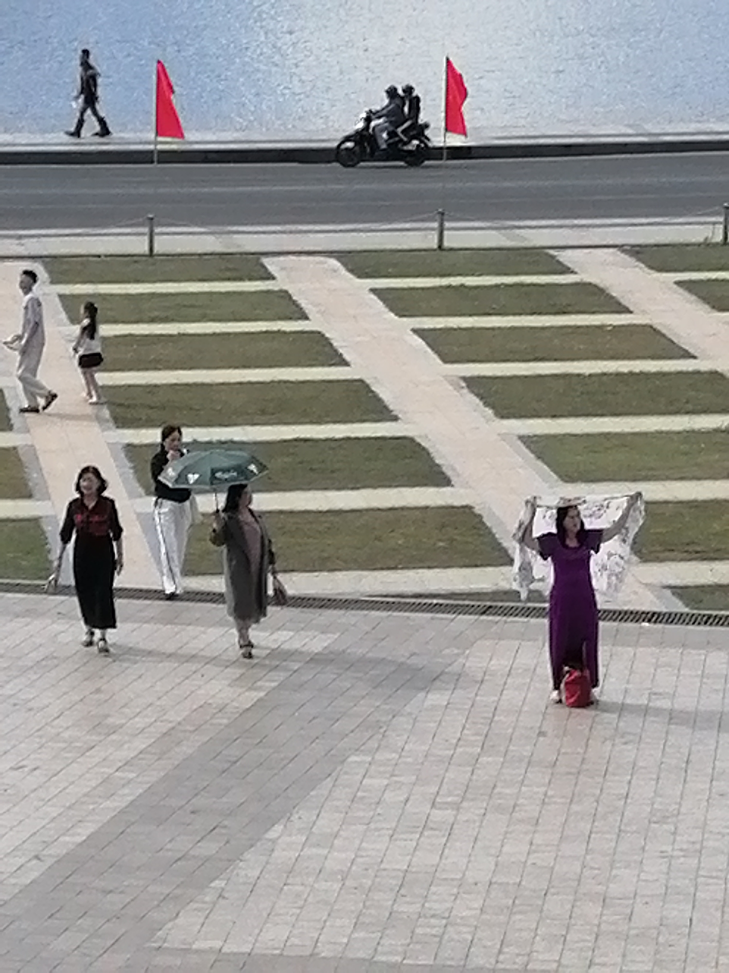 This image depicts a wide, paved outdoor area adjacent to a body of water, possibly a lake or reservoir. In the background, a man walks along a road, and two people are on a motorcycle. A red flag is planted near the road. In the middle ground, a person in a light-colored outfit walks with a child. Further forward, three women are walking. One woman, in a black dress with red embroidery, is in the foreground. To her right, another woman in white pants and a dark top walks with a green umbrella. To the right of the woman with the umbrella, a woman in a purple dress holds up a white, lace-like garment. A red bag is on the ground near her feet. The paving stones are arranged in a patterned walkway. The area appears to be a public space in Da Lat, Vietnam.