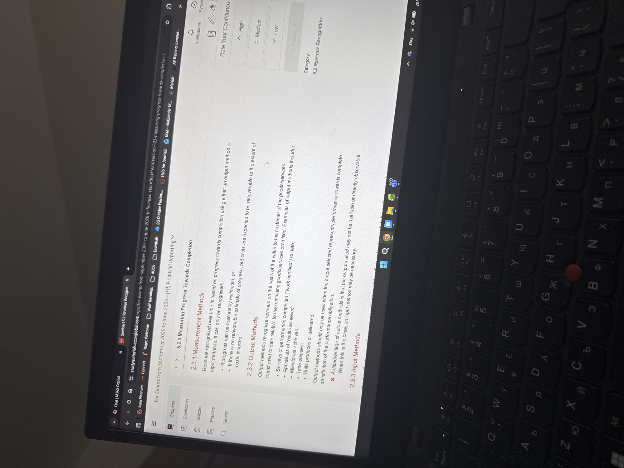 A photograph shows a laptop screen displaying study materials for an ACCA Financial Reporting exam, specifically Section 5.2 Revenue Recognition. The title on the webpage reads "5.2.3 Measuring Progress Towards Completion". The page discusses measurement methods, including output and input methods. Text indicates the materials are for exams from September 2025 to June 2026. The laptop's keyboard is visible, showing Cyrillic letters. The laptop is black, and the screen also shows a "Rate Your Confidence" feedback form with "Medium" selected. There are notifications and a synced status indicator in the upper-right corner of the screen. The environment appears to be indoors, likely a home or office, given the presence of the keyboard. The location is not explicitly stated, but the presence of Cyrillic letters on the keyboard hints towards a Slavic country, specifically it could be Bulgaria since that the Cyrillic alphabet is used in Sofia, Bulgaria.