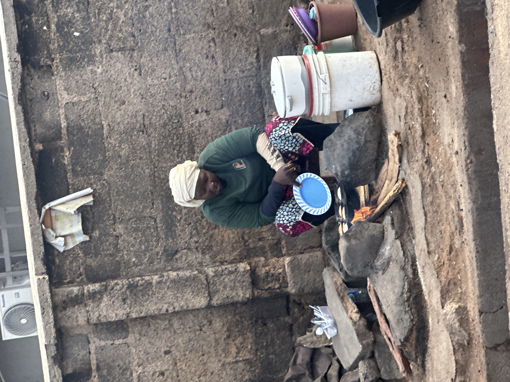 A woman is squatting next to a fire pit in what appears to be an outdoor cooking area. She is wearing a light-colored headscarf and a green shirt, with a patterned skirt. In front of her, she holds a blue plastic plate. Beside her, there are several containers, including a white bucket with a lid and a brown bucket with colorful items inside. The background is a rough, stone wall. Scattered around the cooking area are rocks, pieces of wood for the fire, and some plastic utensils in a container. The ground is dirt and gravel. This scene is located in Gura Top, Nigeria.