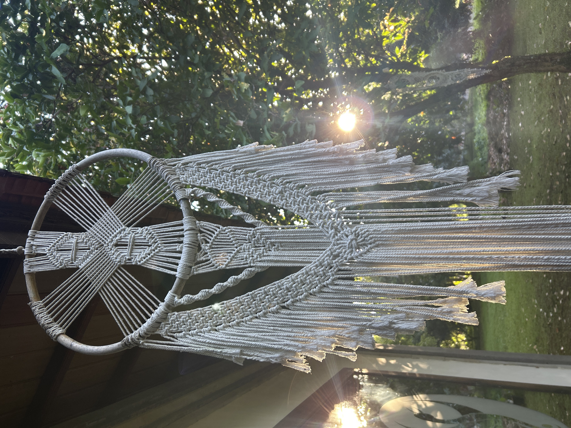 The image shows a macrame wall hanging displayed outside. The macrame is primarily white or off-white and features a circular frame with intricate woven patterns within. Strings hang down from the frame. The background includes green trees, and sunlight filters through the leaves, creating a lens flare effect. Part of a building structure is visible on the left side of the frame, with a roofline. The reflection of the sun and leaves is visible on the lower part of the picture, which might be a window. The precise location within the city of Kediri, Indonesia, cannot be determined from the image.