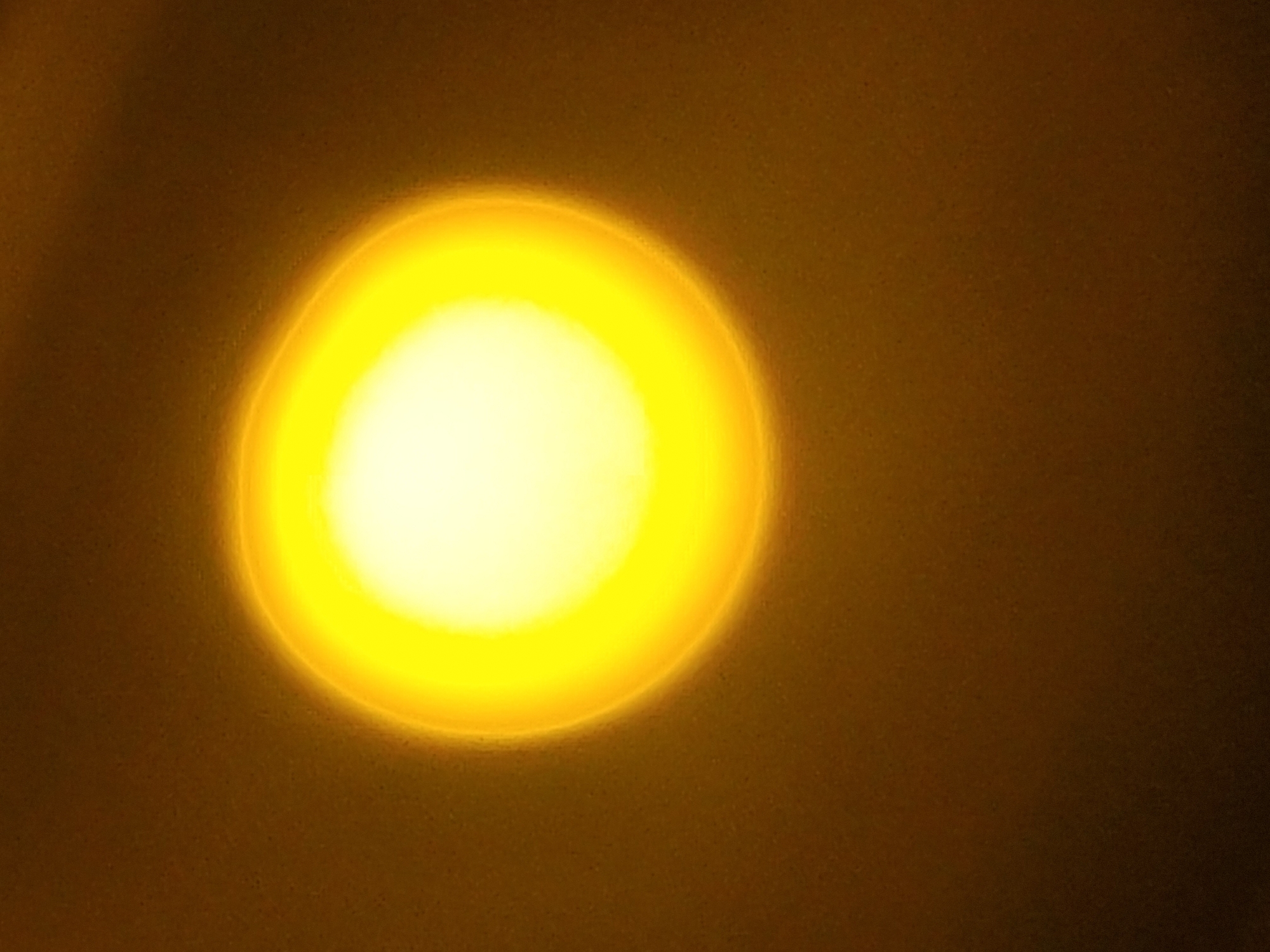 A blurred, circular, bright yellow light dominates the left-center of a textured, dark yellow-brown background. The light is brightest in the center, fading to a soft yellow ring around the perimeter. The background is darker towards the right edge of the frame. The overall texture suggests a close-up of a wall or similar surface. Location: Unknown, Unknown.