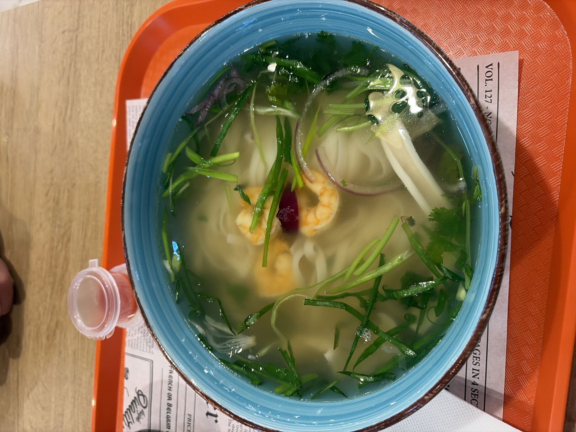 An overhead shot captures a bowl of Asian-style noodle soup, likely pho, served in Izhevsk, Russia. The light blue ceramic bowl with a brown rim holds a clear broth with white flat noodles, several cooked shrimp, thin slices of red onion, and generous portions of chopped green onions and cilantro. The bowl rests on a white paper liner, which in turn sits on an orange, textured plastic tray. A small, clear plastic cup containing a red condiment, possibly chili sauce, is positioned to the left of the bowl on the tray. The underlying table surface is light wood. The paper liner features partially visible text, resembling a newspaper or magazine, with phrases like "VOL. 127 - NG", "PAGES IN 4 SEC", "PRICE", "FRENCH OR BELGIUM", and "Qualit" discernable. The scene depicts a ready-to-eat meal in a casual dining environment.