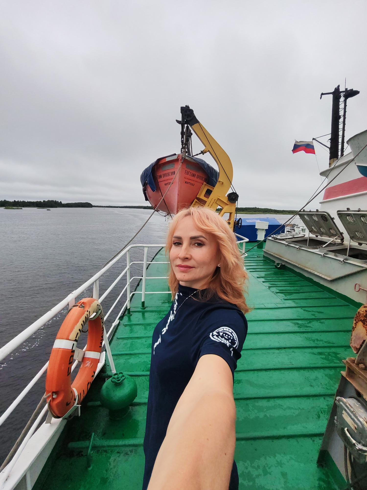 A fair-skinned woman with shoulder-length blonde hair and light makeup takes a selfie on the deck of a ship under a cloudy sky. She is wearing a dark blue t-shirt with a circular emblem on the left sleeve. To her left, a bright orange life preserver hangs on a white railing. Beyond the railing is a body of water with a distant tree-lined shore. A red life boat is suspended from a yellow crane overhead. A Russian flag flies from a mast on the ship behind her. The ship's deck is painted green.