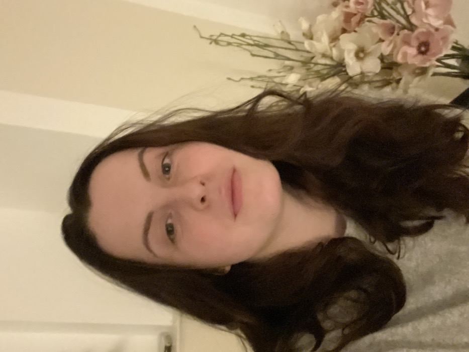 A woman with long, dark brown hair gazes directly at the camera. She has fair skin, light eyes, and a subtle smile. A bouquet of white and pink flowers sits behind her head, partially blurred. The background appears to be a light-colored wall, and she is wearing a light gray sweater. The image is slightly rotated.