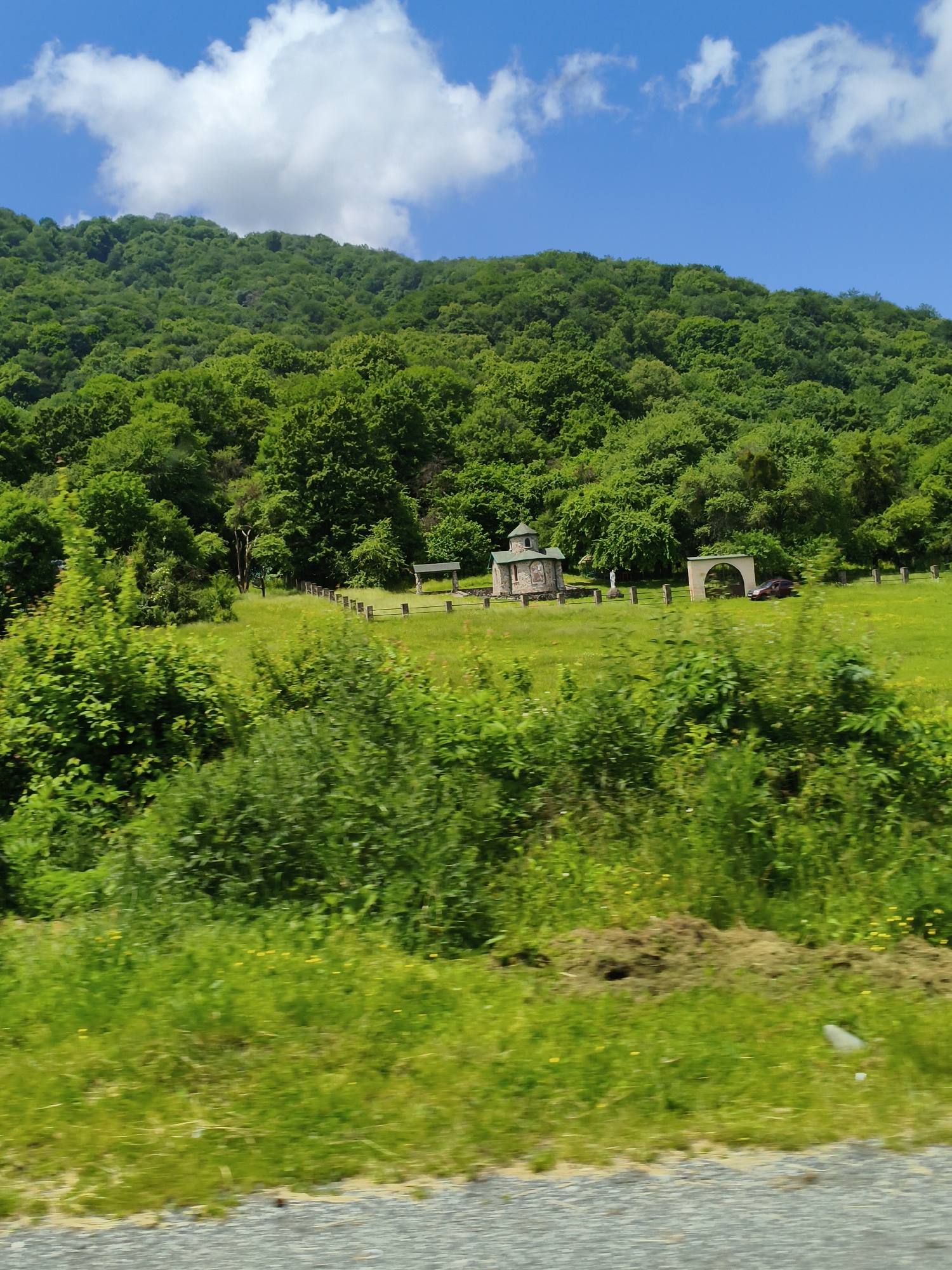 The image depicts a serene, rural landscape under a bright blue sky dotted with fluffy white clouds. Lush, green trees blanket a rolling hillside that dominates the background. In the mid-ground, a vibrant green meadow stretches out, bordered by a low wooden fence. Several man-made structures are scattered across this field. To the left, a small, stone church with a domed roof stands prominently. Next to it, a statue of a person is visible. Further to the right, an arched gateway frames a passing car. A picnic table or shelter is also present in the field. The foreground is filled with more dense, green foliage, appearing somewhat blurry, suggesting the photo was taken from a moving vehicle. The overall impression is one of natural beauty and tranquility, possibly a remote, religious site in a mountainous or hilly region. The bright daylight and clear sky indicate fair weather. There is no visible text in the image.