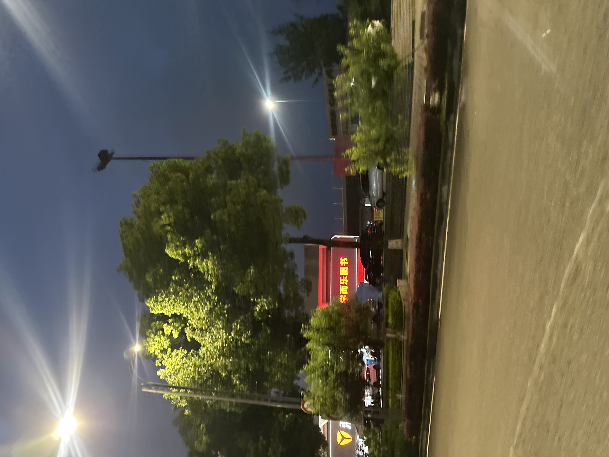 The image depicts a street scene at dusk or night in the Chongqing High-tech Zone, China. A bright street light illuminates the sky and the upper branches of a large, leafy green tree. In the background, a red building with illuminated signage is visible, with the characters "学而乐图书" (Xue Er Le Tu Shu), meaning "Learn and Happy Books," prominently displayed. Several vehicles, including cars, are parked or driving on the street. The foreground is dominated by a road surface. The overall atmosphere is urban and illuminated by artificial lights.
