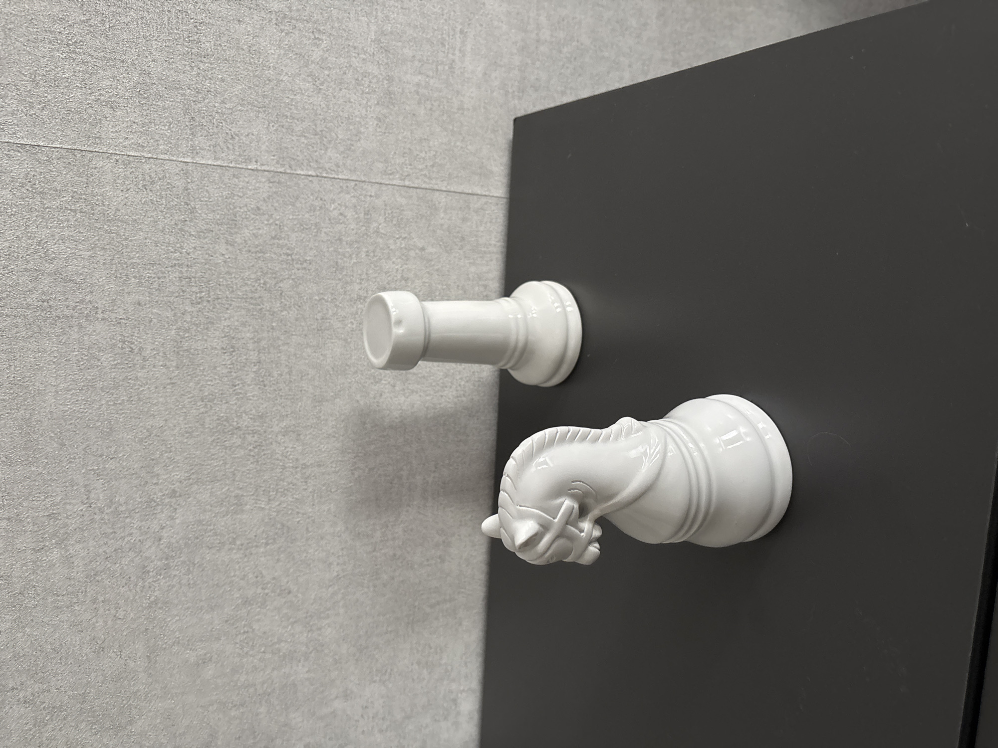 Two white ceramic chess pieces, a knight and a rook, are displayed on a dark, flat surface. The knight is positioned in the foreground on the left, facing left, while the rook is in the background on the right, facing forward. Behind the chess pieces, a textured gray wall is visible. The lighting in the scene is even, casting subtle shadows on the surface. The overall setting appears to be an indoor environment, possibly a home or a display area.