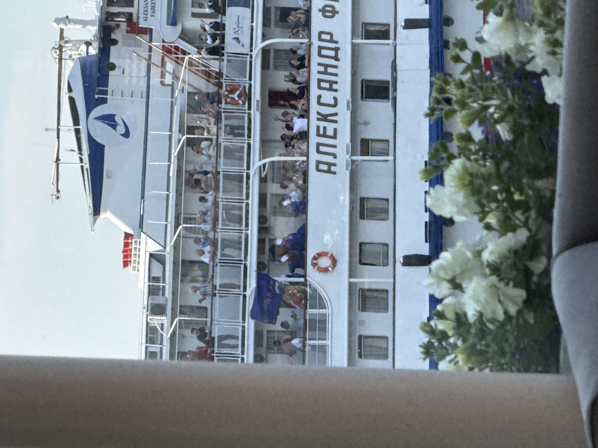 The image shows a white cruise ship named "АЛЕКСАНДР ФАДЕЕВ" docked in the city of Perm, Russia. The ship has multiple decks with people standing on the railings, some waving. A blue flag is visible on one of the decks. In the foreground, there are white flowers and greenery, suggesting a waterside location. The sky is clear and light blue. The ship's name is prominently displayed in large Cyrillic letters on its side. A logo with a blue sailboat is visible on the upper deck. The overall scene depicts a lively atmosphere on the ship, possibly during a departure or arrival.