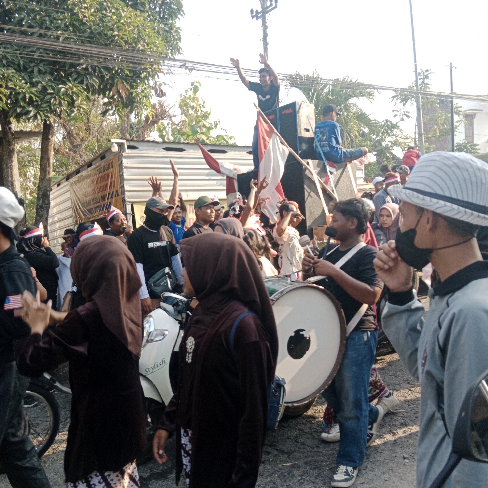 The image depicts a street scene in Tegalrejo, Indonesia, under bright daylight. A group of people are gathered, with several individuals actively participating in what appears to be a procession or celebration. In the background, a truck or trailer is visible, serving as a makeshift stage. On top of this platform, a young man in a black t-shirt with his arms raised is enthusiastically cheering, positioned near large speakers and a flag. Another individual in a blue shirt is seated on the platform.
In the foreground and mid-ground, there is a diverse crowd. Some individuals are wearing head coverings, including hijabs. A person in the center is playing a large drum, holding drumsticks. Another person to the right, wearing a light grey long-sleeved shirt and a fedora-style hat, has a black mask covering their face and is looking to the side. Several people are holding Indonesian flags, red and white, some of which are attached to poles. There are also glimpses of motorcycles and what appears to be a banner with text on the side of the trailer. The ground is asphalt, and trees line the street, with power lines running overhead. The overall atmosphere suggests a lively gathering, possibly a parade or a public event.