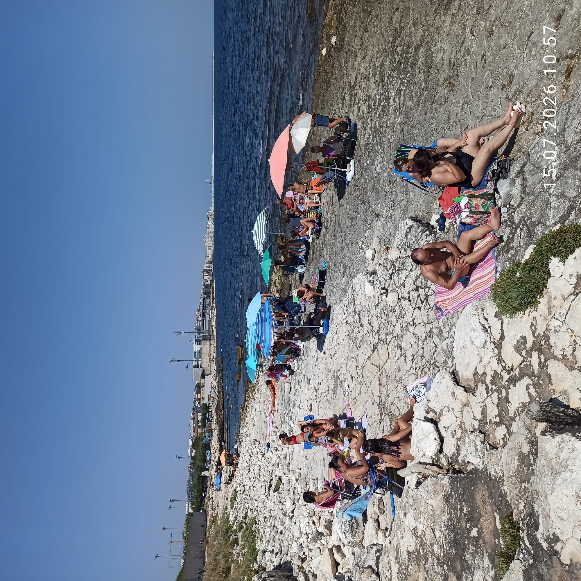 In Molfetta, Italy, a rocky coastline is dotted with sunbathers on a clear, sunny day. The sea is calm and blue. People are spread out across the rocks, some sitting in chairs, others on towels, and a few are in the water. Beach umbrellas provide shade for some of the groups. In the background, the city of Molfetta is visible with its buildings and a harbor. The overall scene depicts a typical summer day at the beach.