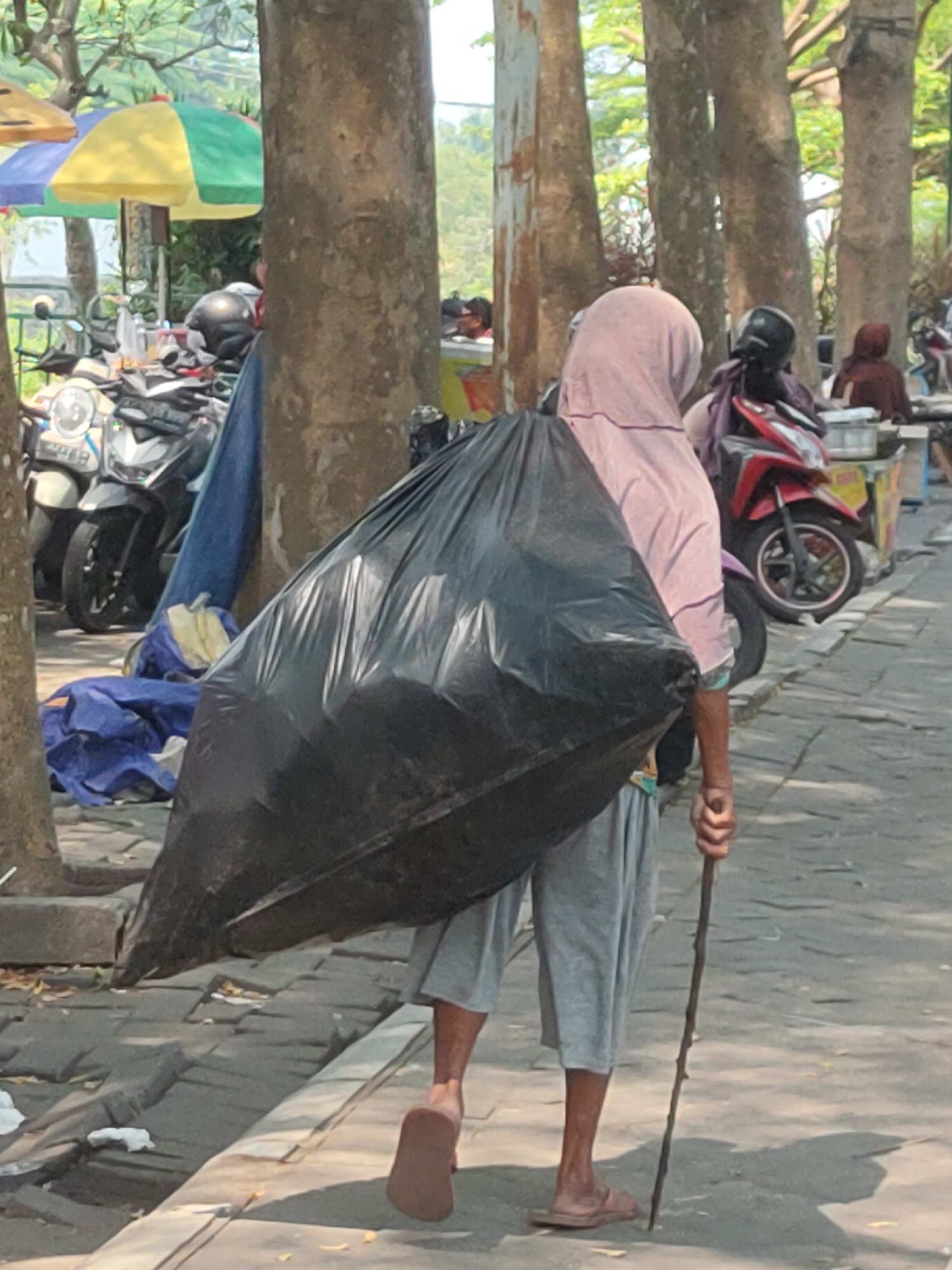 In the city of Ngasem, Indonesia, a person wearing a pink headscarf and a light pink shirt walks on a stone-paved sidewalk, carrying a large black trash bag slung over their left shoulder. They are also using a long, thin stick as a walking aid. The trash bag appears to be full and is positioned behind the person, obscuring their back. The person is wearing grey shorts and brown sandals. In the background, several motorcycles are parked, and some colorful umbrellas are visible, suggesting a market or street vendor area. Trees with rough bark line the sidewalk, casting shadows on the ground. The overall scene depicts a person engaged in collecting or transporting refuse in an urban environment.