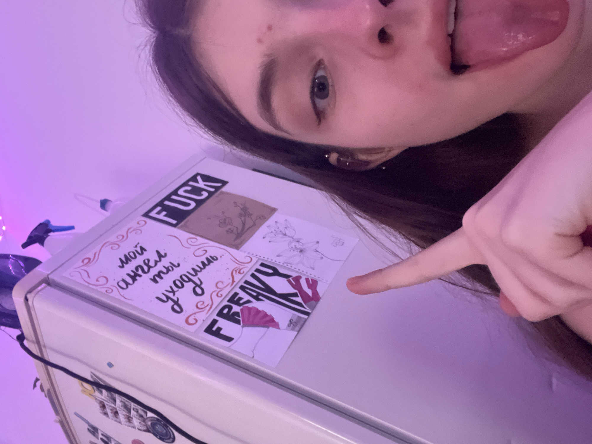 A young woman with brown hair and an earring is pointing at a white refrigerator door. The refrigerator has multiple papers stuck to it. The papers are adorned with various sketches, phrases, and words, including "FUCK," "FREAKY," and "мой ангел ты уходишь" (my angel, you are leaving). The scene appears to be indoors, with a purple light illuminating the background. The location is not apparent in the image.