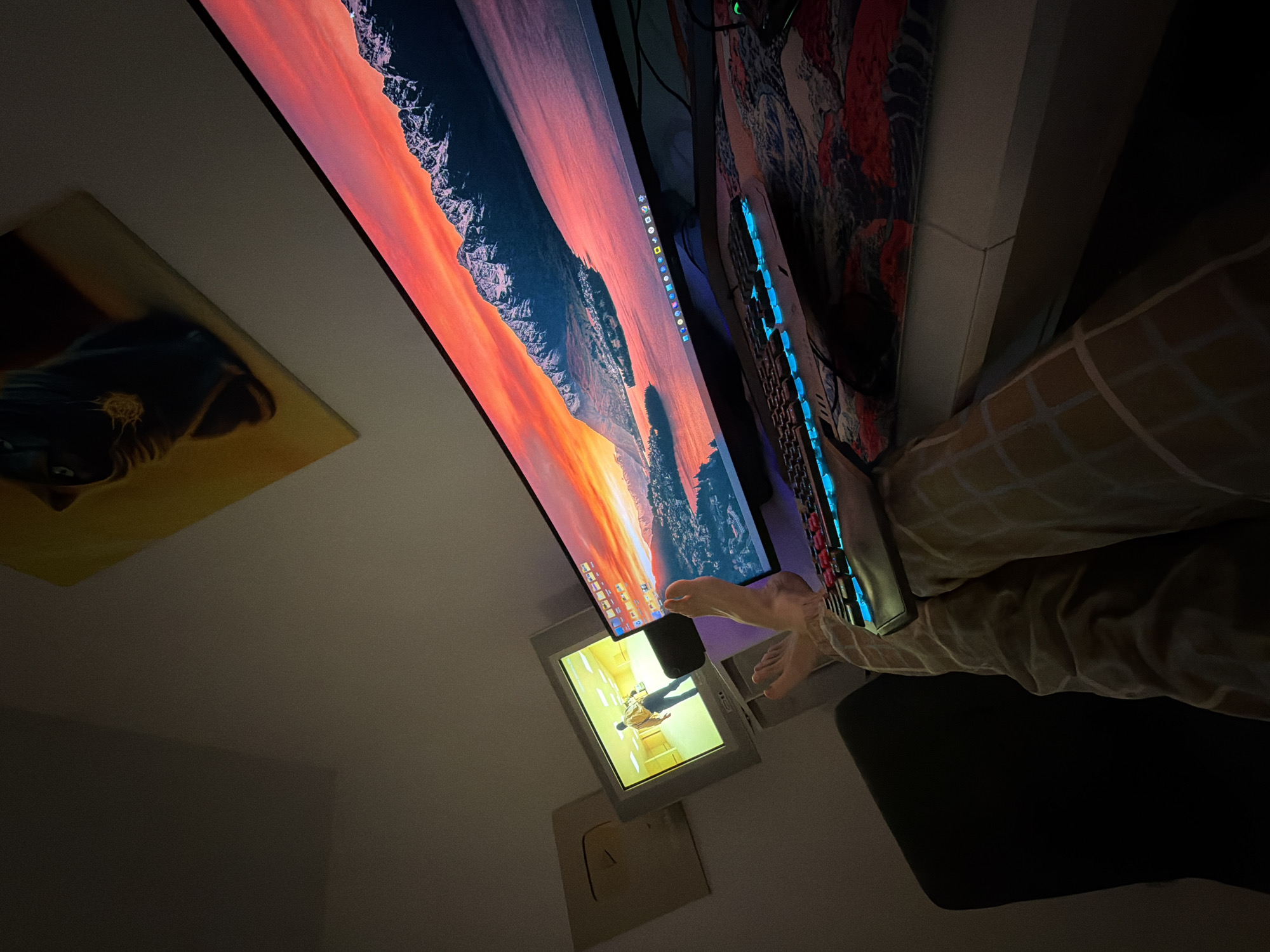 The image shows a person's feet and legs resting on a desk in front of a computer setup. A large, curved monitor displays a vibrant sunset over a mountainous landscape. To the left of the monitor, a smaller, older television displays a yellow-toned image of a person walking in a room. A gold-colored plaque, resembling a YouTube play button, is mounted on the wall behind the smaller television. The desk is covered with a patterned cloth and a glowing keyboard. The overall environment suggests a home office or entertainment space. There is no information in the image to suggest the location is Naberezhnye Chelny, Russia.
