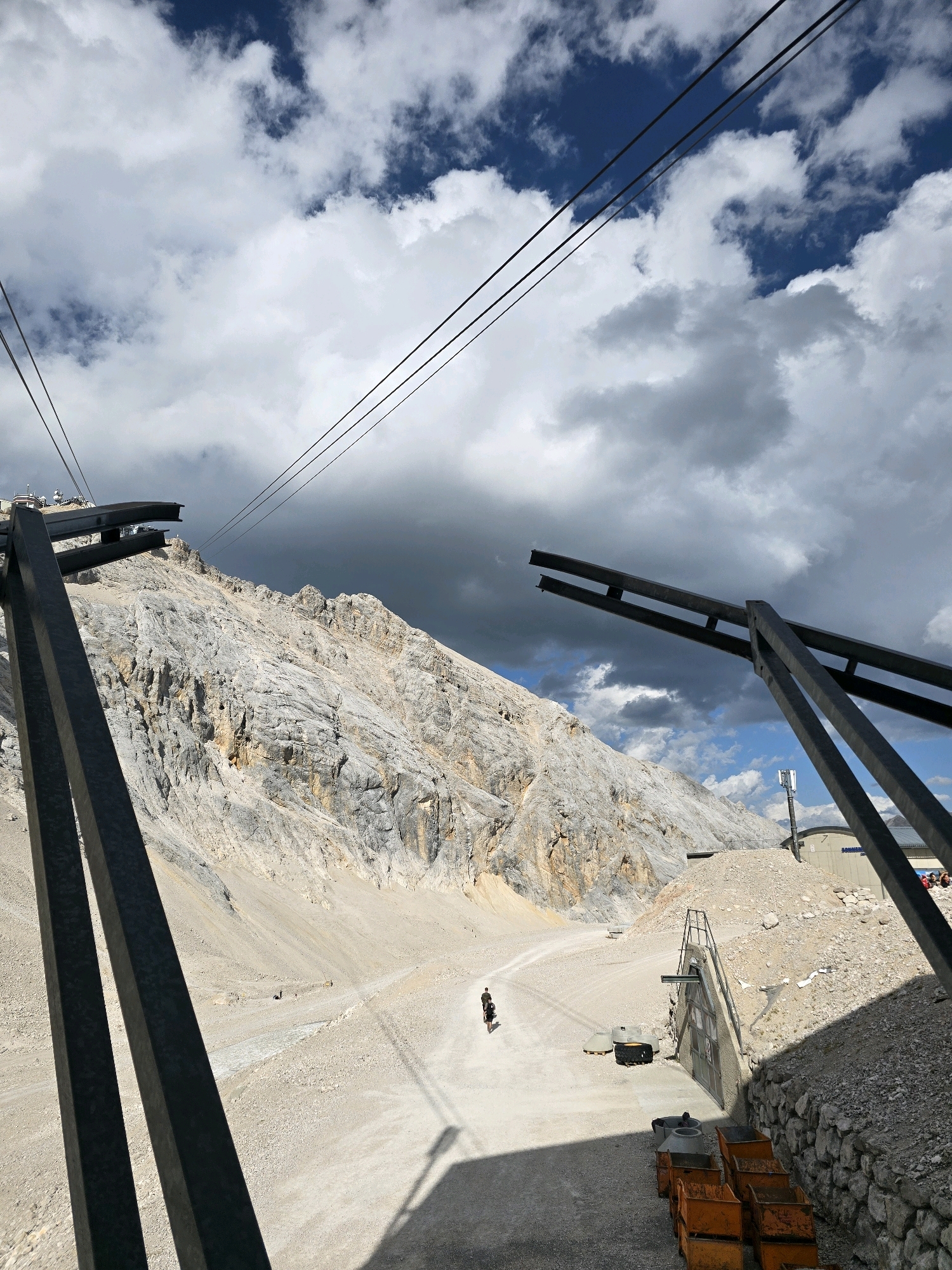 The image depicts a mountain landscape, likely taken from a cable car station or a viewpoint near the Zugspitze, Germany's highest peak, which is accessible from Garmisch-Partenkirchen. In the foreground, large dark metal structures, possibly part of a cable car system, dominate the view. The mountain slopes are rocky and barren, with sparse vegetation. A dirt path winds through the lower part of the scene, where a lone cyclist is visible, casting a shadow on the ground. In the middle ground, there is a building with an entrance, and a communication tower stands tall. The sky is dramatic, with a mix of dark storm clouds and patches of blue sky. The overall impression is of a rugged, alpine environment under a dynamic sky.