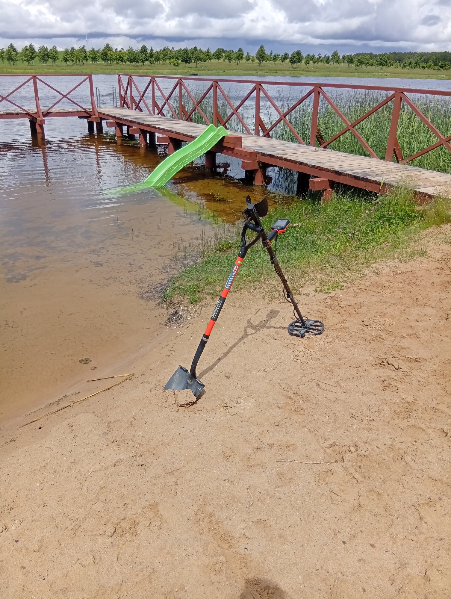 The image depicts a sandy shoreline adjacent to a body of water, likely a lake, in Šaukėnai, Lithuania. A wooden boardwalk with a red railing extends over the water. A bright green slide is partially submerged in the water, attached to the boardwalk. On the sandy shore, a metal detector stands upright, leaning against itself. A shovel with a black handle and a metal blade is placed in the sand near the water's edge. The background shows a treeline and a cloudy sky with wind turbines visible in the distance. The water is calm and appears shallow near the shore, with visible sandy bottom and some vegetation.
