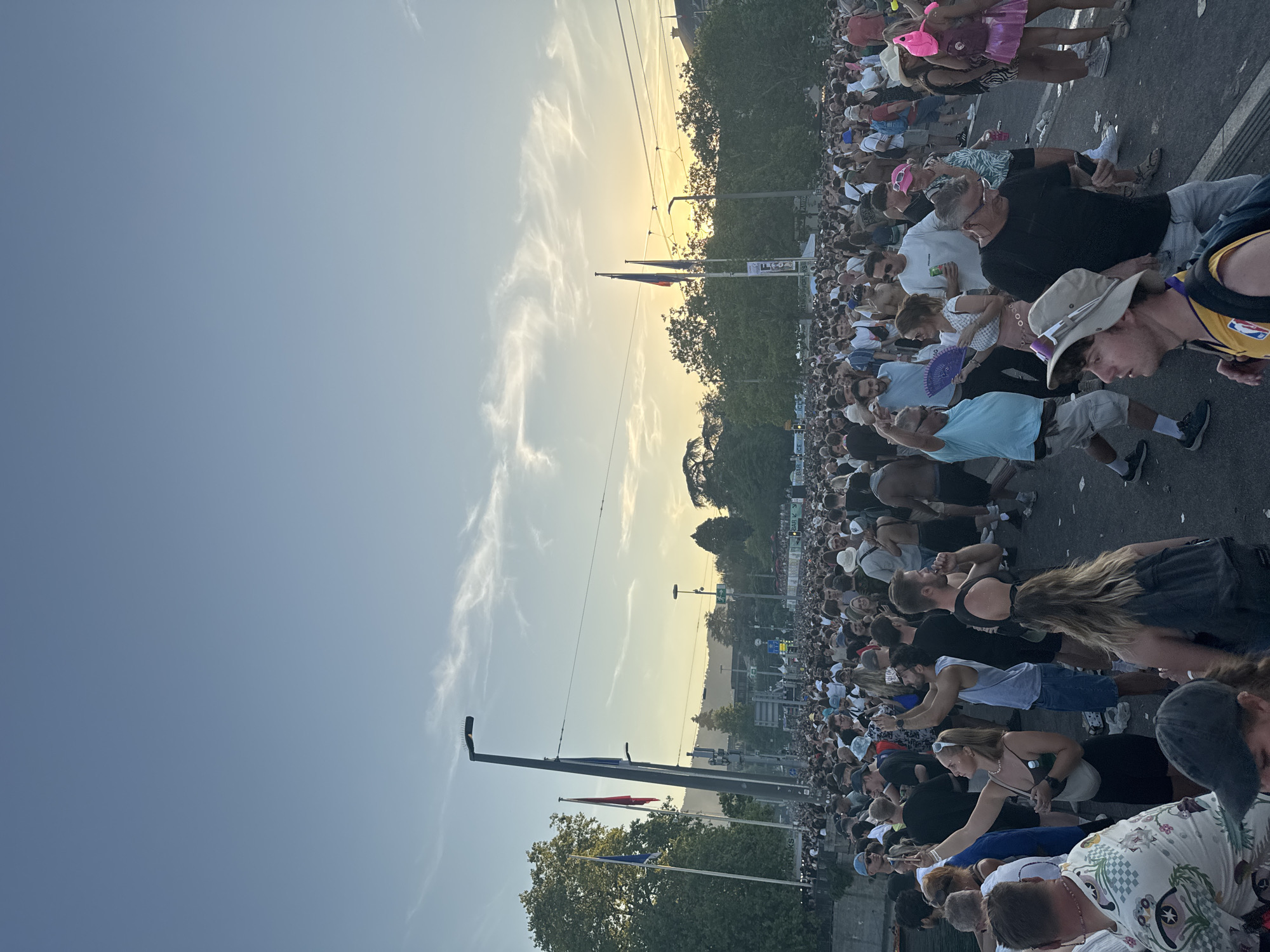 The image depicts a densely populated street scene in Zurich, Switzerland, during what appears to be a daytime event or festival, with the sun setting or low in the sky. A large crowd of people, dressed in casual summer attire, fills the width of the street. Many individuals are captured in candid moments, with some taking photos or videos with their phones, dancing, or conversing. Several streetlights and flags are visible, along with trees lining the background. The overall atmosphere suggests a lively and crowded public gathering.