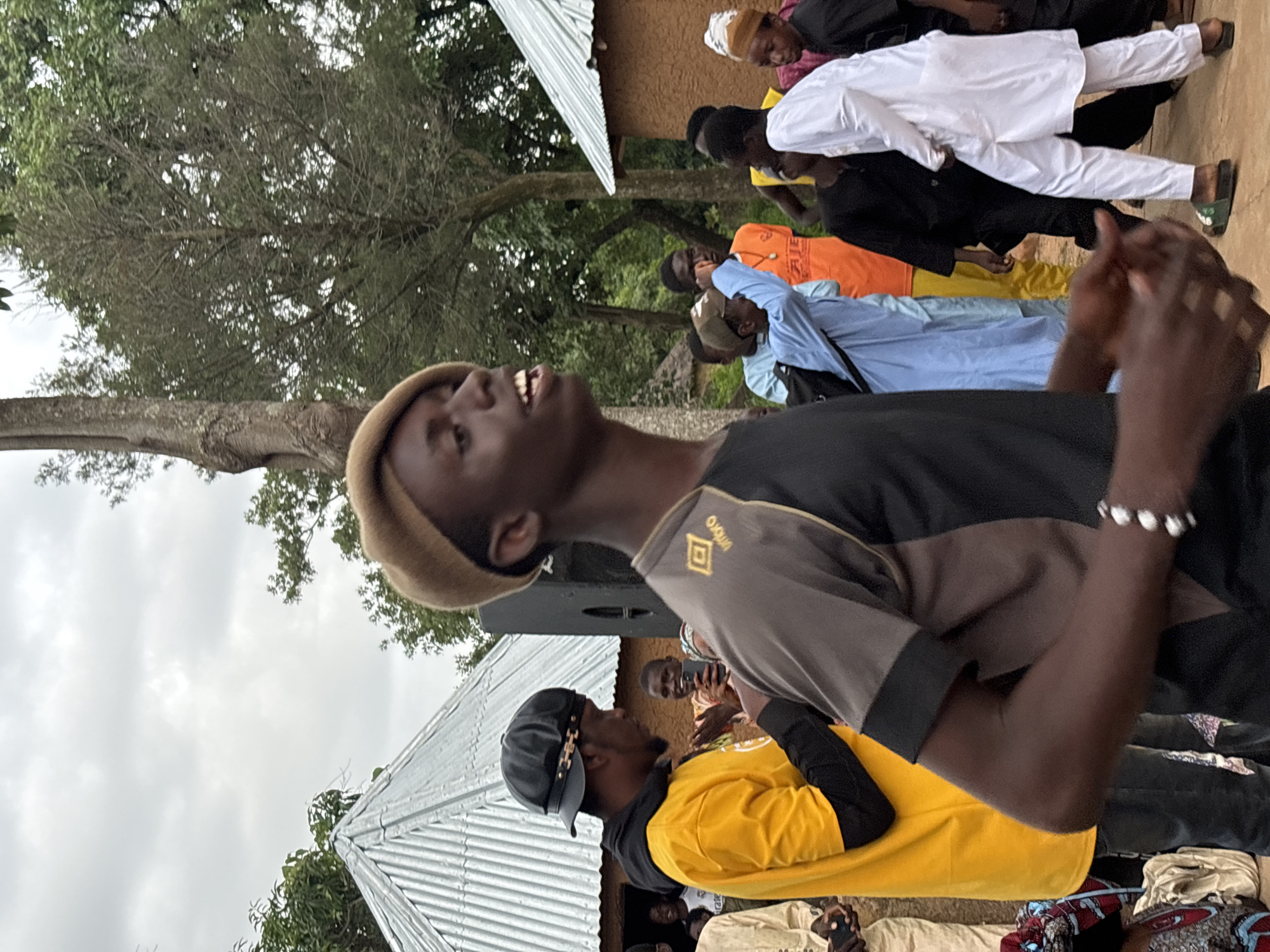 The image depicts an outdoor gathering of people, likely in a community setting in Jos, Nigeria. The foreground is dominated by a young Black male wearing a tan beret and a grey and black t-shirt with a gold logo. He is looking to his right with his mouth open, suggesting he might be speaking or singing. His hands are clasped together in front of him, and he wears a bracelet on his left wrist.
Behind him, other individuals are visible. To the left, a Black male in a bright yellow hooded jacket and black pants is looking down. Further in the background, a group of people are gathered. One individual in an orange t-shirt stands out. Another person in a light blue garment is gesturing with their hand. Several others are dressed in traditional attire, including one person in a white robe and another in a dark outfit.
The environment suggests a rural or semi-rural area with traditional housing structures visible. One building has a corrugated metal roof, while another appears to be made of mud or adobe. Trees and foliage are present in the background, indicating a natural setting. The sky is overcast, suggesting a cloudy day. The overall scene conveys a sense of community event or social gathering.