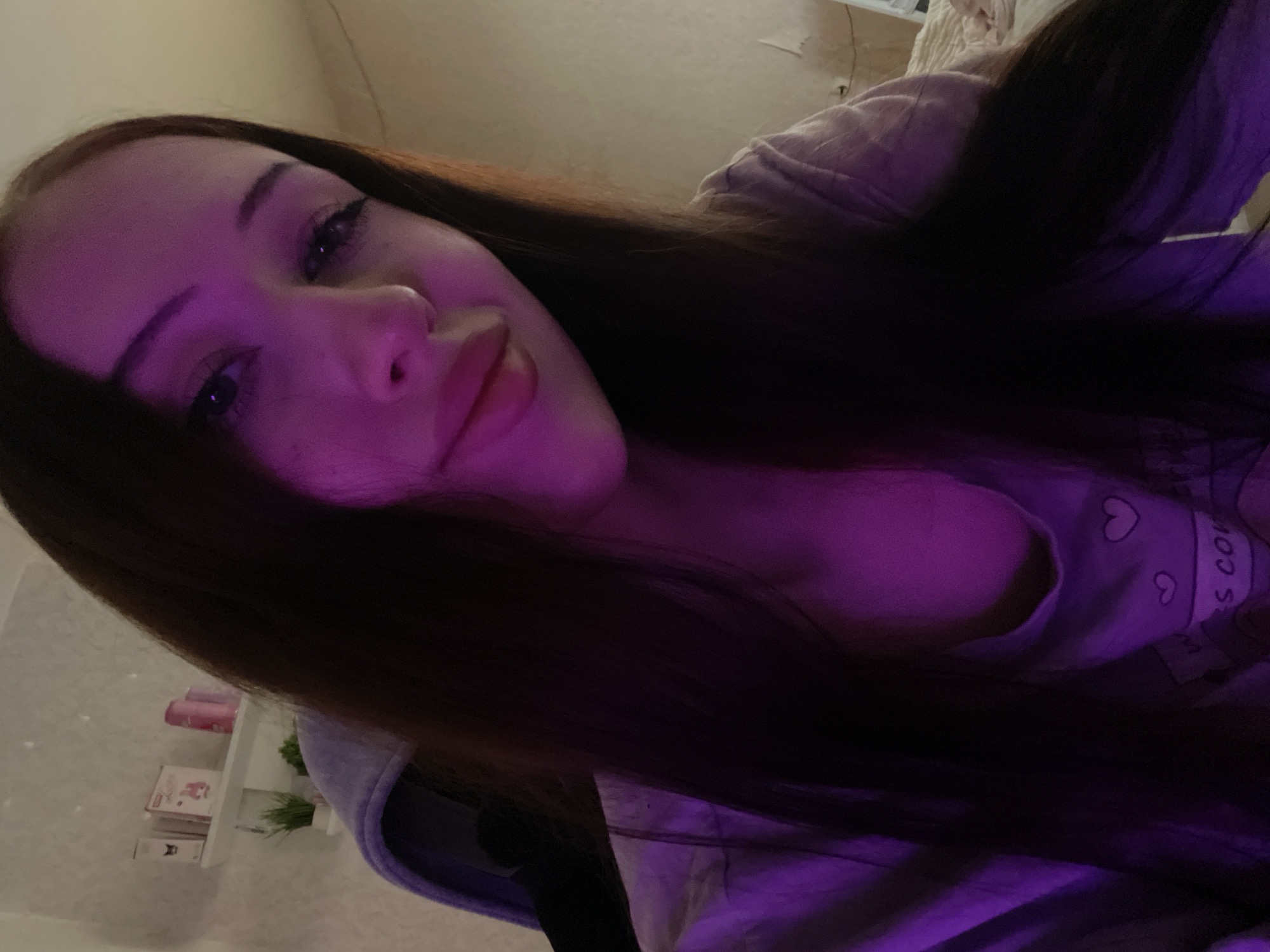 A young woman with long dark hair and fair skin is captured in a selfie, angled from above. She is wearing a light purple hoodie with hearts and text that reads "S CO." The image is illuminated by purple light, suggesting it may have been taken in the evening or with artificial lighting. Her expression is relaxed, with her lips slightly parted. The background is blurry, but appears to be an indoor setting, possibly a bedroom, with shelves holding some small items. The overall mood is casual and intimate.