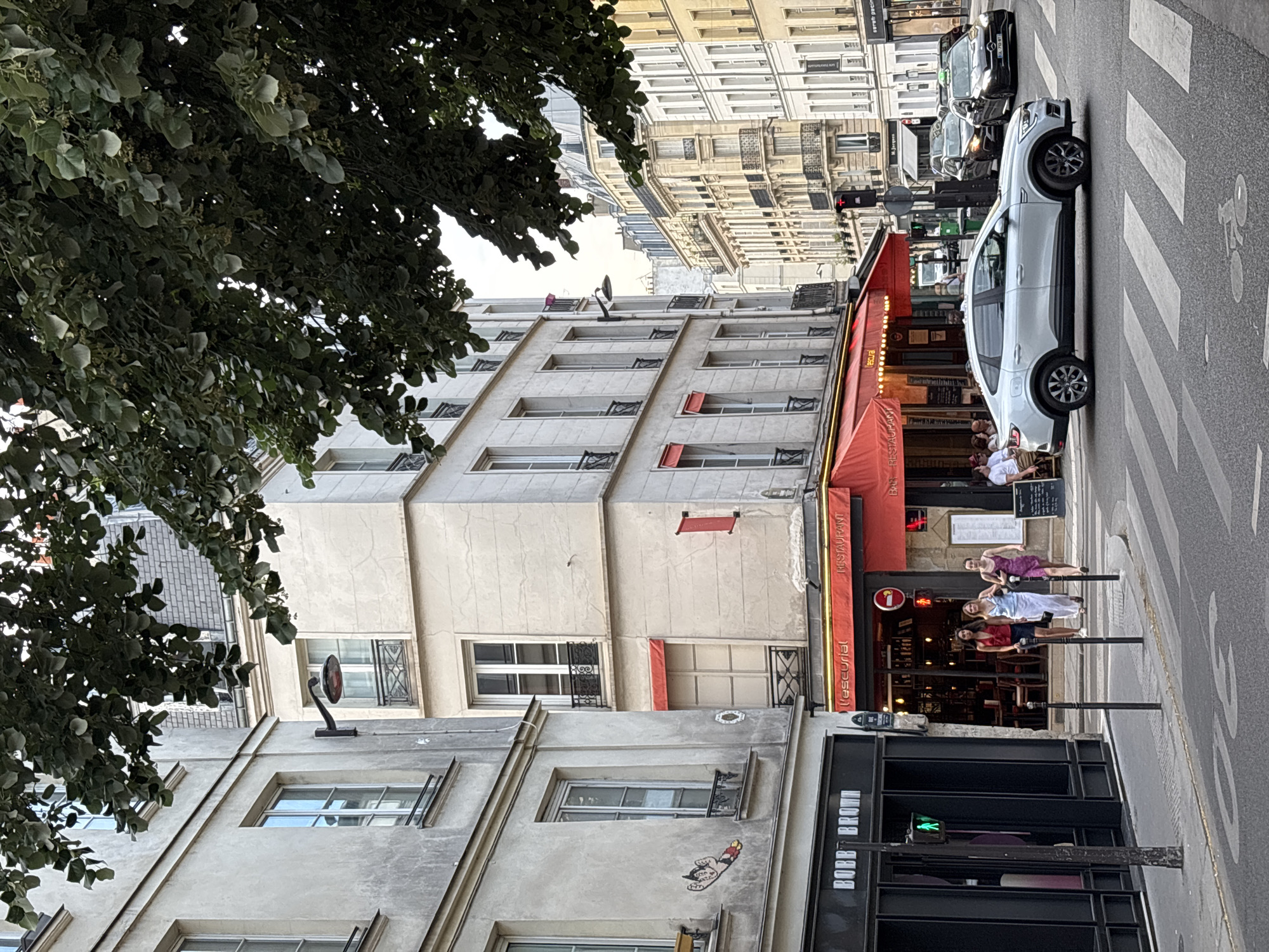 This is a street scene in Paris, France, focusing on a corner intersection with multiple buildings and pedestrian activity. On the left, a light-colored building has a black storefront with "BOBB BROWN" signage and a green pedestrian crossing signal. Adjacent to it, a building with a red awning and "l'escurial RESTAURANT" signage is visible. Three women are walking across the street in front of the restaurant. Further down the street, several cars are present, including a light-colored sedan crossing the intersection and two dark-colored sedans approaching. The architecture is characteristic of Paris, with multi-story buildings featuring balconies and dormer windows. Overhead, leafy green branches from a tree partially obscure the upper portions of the buildings. The overall impression is a bustling urban environment in daylight.