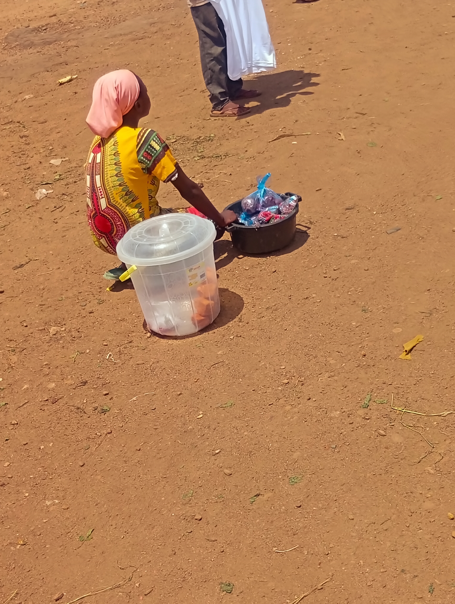 A person wearing a yellow and red patterned dress and a pink headscarf sits on the ground in Bukuru, Nigeria. In front of them is a large, clear plastic container with a lid, and to their right is a black bucket filled with packaged items, some in blue plastic bags. The ground is dry and reddish-brown dirt, with some scattered debris. Another person, partially visible in the background, stands wearing dark pants and holding a white cloth.