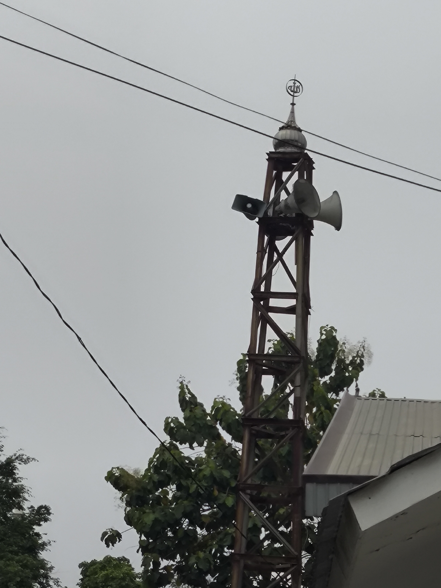 In the city of Cilegon, Indonesia, a tall, weathered metal tower stands against a cloudy sky. Atop the tower is a decorative Islamic crescent moon finial with Arabic script, mounted on a domed structure. Two large loudspeakers, one pointing left and the other to the right, are affixed to the upper section of the tower. Below the speakers, a lattice of rusted metal beams forms the tower's structure. In the background, lush green trees with some white blossoms are visible, partially obscuring a light-colored, corrugated metal roof of a building. Power lines crisscross the sky in the upper portion of the image. The scene is captured from a low angle, looking upwards towards the tower and the sky.