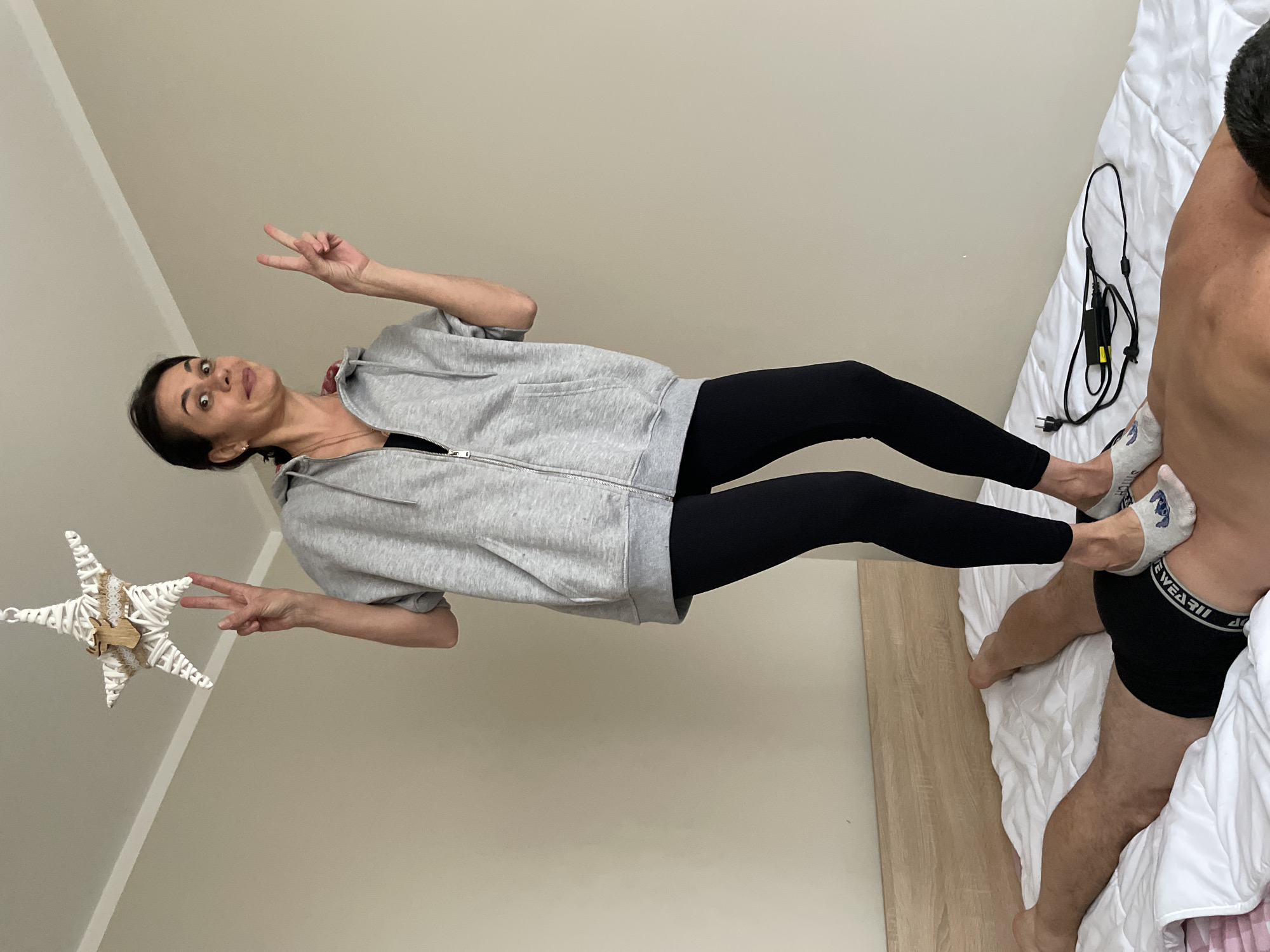 The image depicts an interior bedroom scene. A female subject, wearing a grey zippered hoodie, black leggings, and white socks, stands with both feet on the back of a prone male subject. The female faces the camera with a wide-eyed, open-mouthed expression and displays a peace sign with each hand. The male subject is lying face down on a white quilted comforter on a bed, wearing black boxer briefs, with his back exposed. A light wood headboard is positioned behind the bed. A black laptop charger rests on the comforter near the male's lower back. A white woven star ornament, decorated with a wooden deer head, hangs from the white ceiling. The room walls are painted a light beige color.