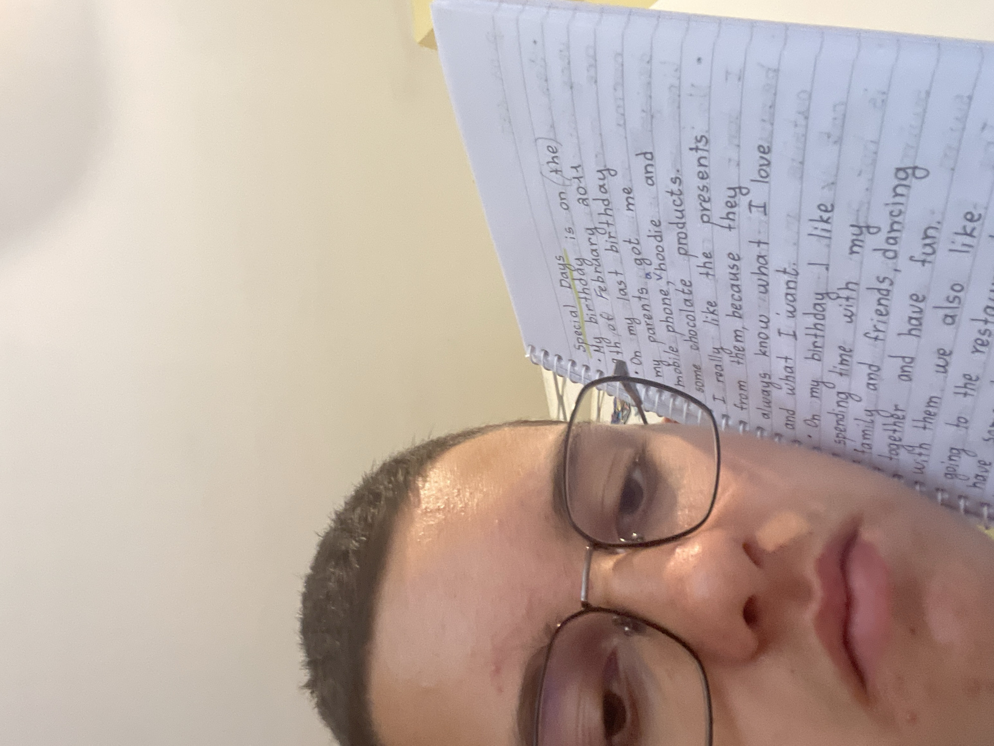 An upright, selfie-style close-up features a person holding a spiral-bound notebook in front of their face, partially obscuring it. The person, whose gender is not explicitly discernible, has short, dark hair and wears dark-framed glasses. Their eyes are visible above the notebook, looking towards the viewer, and a portion of their nose and mouth are visible below the notebook. Their expression appears neutral.
The notebook, held at an angle, displays handwritten text in blue ink on lined paper. The visible text describes "Special Days," with the most prominent sections detailing a past birthday. The text states: "My birthday is on the 9th of February 2011. On my last birthday my parents got me mobile phone, hoodie and some chocolate products. I really like the presents from them, because they always know what I love and what I want. On my birthday I like spending time with my family and friends, dancing together and have fun. with them we also like going to the restau..."
The background behind the person is a plain, light, off-white surface, suggesting an indoor setting. The lighting is soft and even. The overall impression is that of a personal record or school assignment being presented. The location context provided is Sofia, Bulgaria.