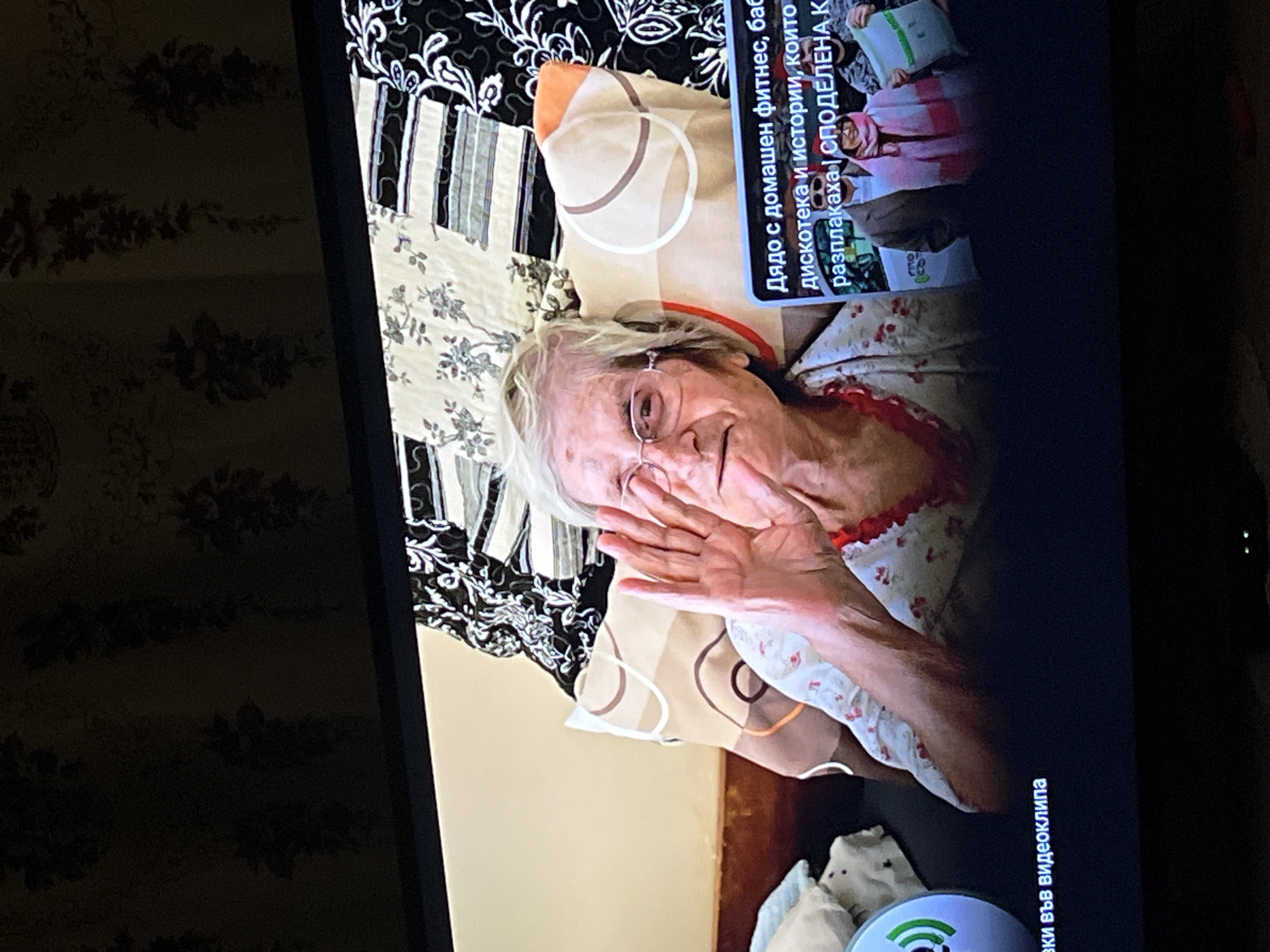 The image shows a television screen displaying a video or broadcast featuring an elderly woman. She is wearing glasses and is lying in bed, waving with her right hand. The bed is made with multiple patterned fabrics, including floral and geometric designs. In the top right corner of the screen, there is text in Cyrillic, along with an image of people, which is part of the video content. The Cyrillic text translates to "Grandfather with home fitness, grandmother disco and stories that made me cry | SHARED K". At the bottom right of the screen, there is more Cyrillic text that translates to "Included in the video clip". The environment is the inside of what appears to be a home, with wallpaper visible on the left side of the screen. The location in the city of Lozarevo, Bulgaria cannot be determined from the provided image.