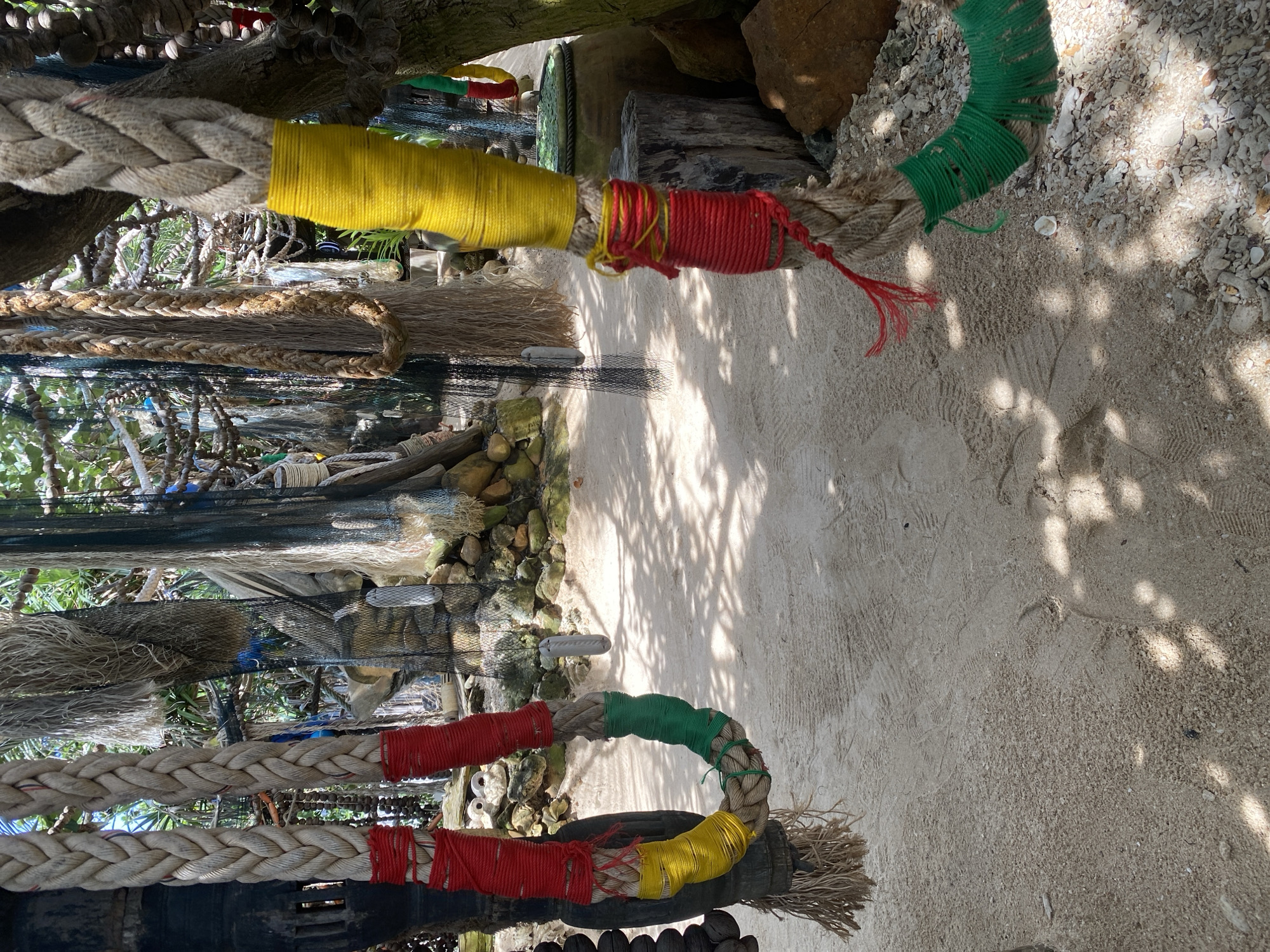 The image depicts a close-up view of thick, braided ropes hanging in an outdoor setting, likely on the island of Koh Phangan, Thailand, given the sandy ground and surrounding vegetation. The ropes are decorative, with sections wrapped in brightly colored yarn: red, yellow, and green. The rope on the left is draped downwards and then loops, showcasing a segment wrapped in red, followed by yellow, and then green. The rope on the right hangs vertically, with a yellow wrap near its upper portion, and a red wrap further down, tied with additional red string. Below the red wrap, a section of the rope is adorned with green fringed material.
The background shows a mix of natural elements and man-made objects. There are trees and foliage, rocks, and sand. Hanging nets of various sizes and textures are also visible, suggesting a coastal or fishing-related theme. The ground is covered in light-colored sand, with some small pebbles and debris. Footprints are visible in the sand, indicating recent activity. Sunlight casts dappled shadows on the ground, suggesting it is daytime. The overall impression is one of a rustic, decorative display, possibly part of a market, art installation, or an establishment in a tropical tourist area.