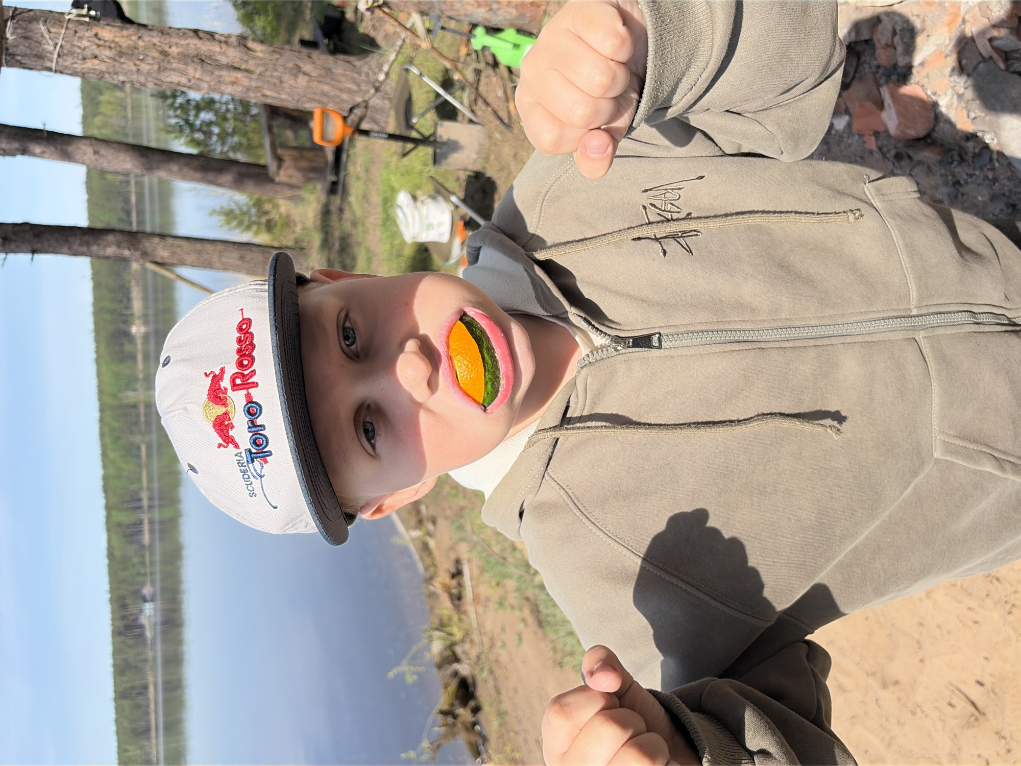 A boy is standing outside near a body of water, wearing a baseball cap and a gray zip-up hoodie. He is holding a slice of fruit in his mouth, possibly an orange or tangerine. His fists are clenched and raised. The background shows trees, the water, and some outdoor equipment. The location is possibly near Забегаево, Russia.