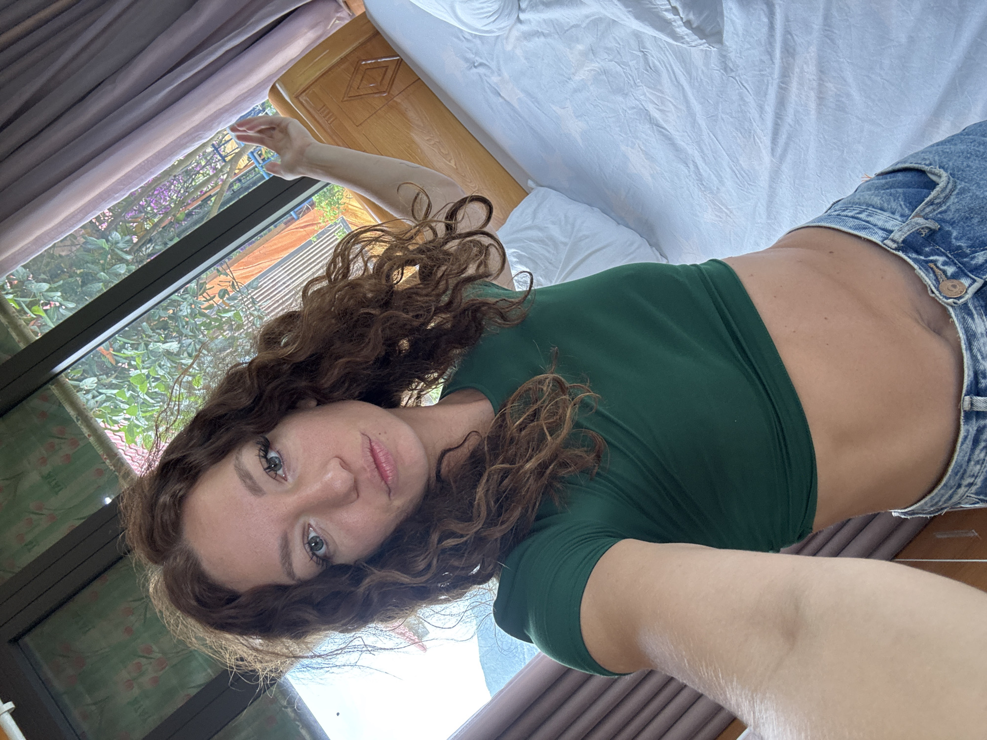 A young, light-skinned woman with curly brown hair and light eyes lies on a bed in Than Uyên, Vietnam. She wears a dark green short-sleeved shirt and blue denim shorts. A window with a view of greenery and purple flowers is visible behind her. The bed has white sheets, and a wooden bed frame is partially visible.