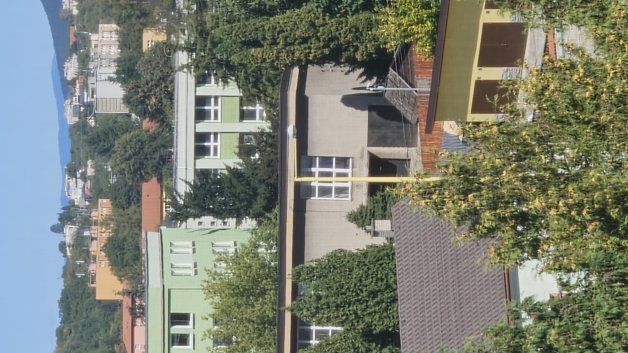 The image depicts a residential area in the city of Prešov, Slovakia, under a clear blue sky. In the foreground, several trees with green foliage and some yellow blossoms partially obscure the view of buildings. A brown-tiled roof of a house is visible on the left. Behind this, a light gray, single-story building with a dark garage door and several windows is present. A yellow pole stands vertically in front of this building, extending upwards towards a street lamp.
Further back, a larger green building with multiple windows is visible. To its right, a light gray building with white-framed windows is seen. In the background, a complex of buildings with orange and light-colored facades, characteristic of Soviet-era architecture, are situated on a tree-covered hill. In the far distance, a range of blue mountains forms the horizon. The overall impression is a moderately dense urban or suburban environment.