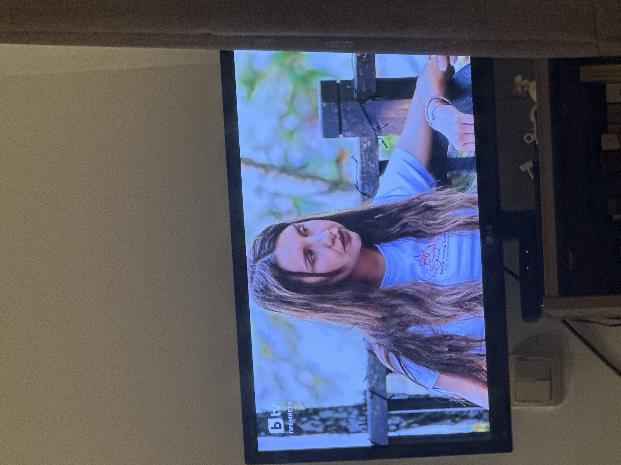 The image shows a television screen displaying a scene with a young woman. She has long brown hair and is wearing a light blue t-shirt. She appears to be sitting on a wooden structure, possibly a bench or a bridge, with a background of blurred greenery suggesting an outdoor environment. The "bTV" logo and the word "премиера" (premiere) are visible at the bottom of the screen, which indicates that the woman is part of Bulgarian television programming. Without further information, it is not possible to determine a specific location in Sofia, Bulgaria.