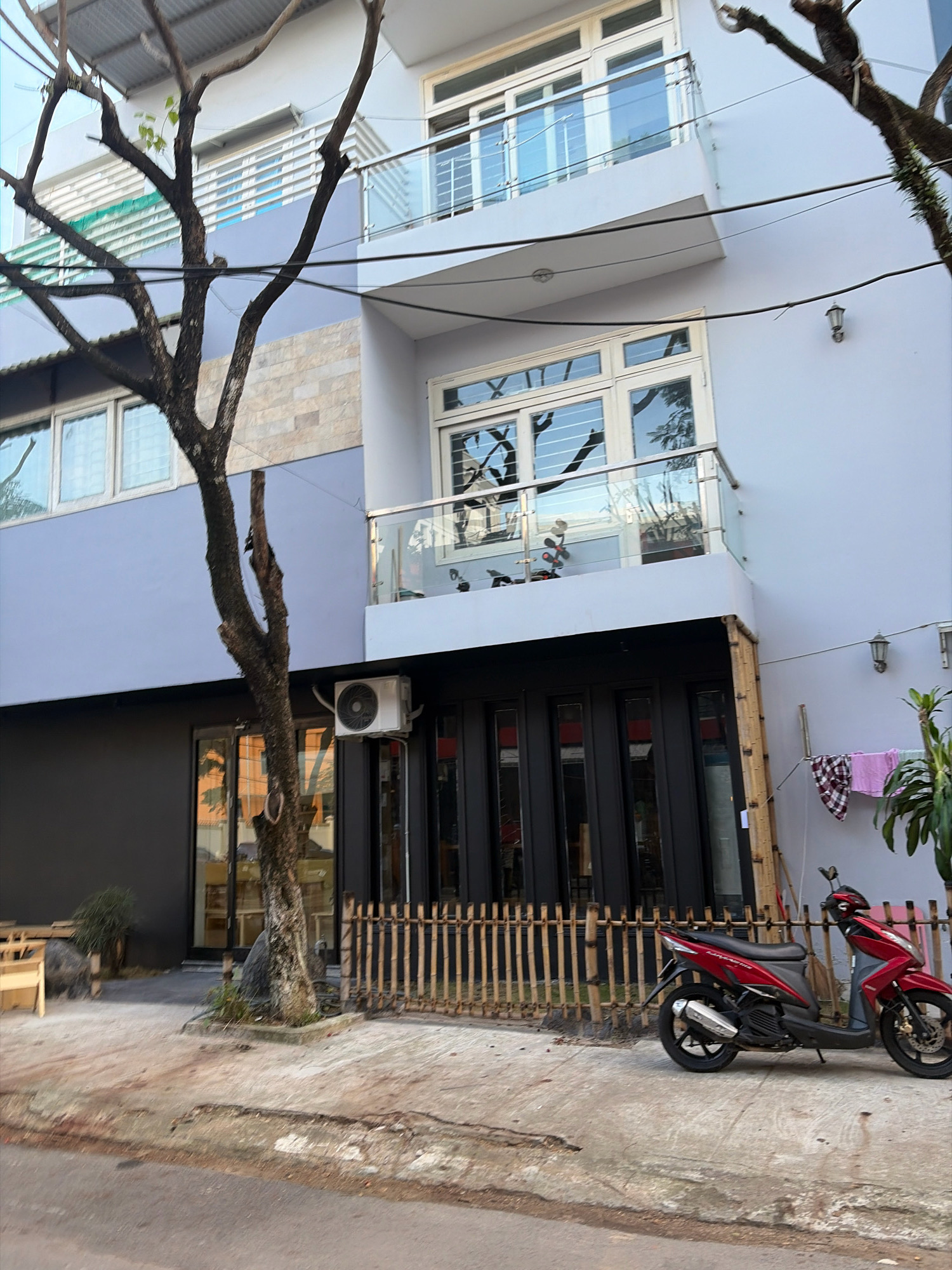 The image depicts the exterior of a multi-story building in the city of Đà Nẵng, Vietnam. The building features a light blue facade on the upper levels and a dark gray facade on the ground floor. Several large windows with white frames are visible on different floors, some with reflective glass showing trees and sky. A balcony with a glass railing extends from the second floor. An outdoor air conditioning unit is mounted on the wall below the second-floor balcony.
In front of the building, a tree with bare branches stands near the sidewalk. A short, rustic fence made of bamboo poles runs along the front of the ground floor. A red scooter is parked on the concrete sidewalk to the right of the fence. Laundry, including a checkered cloth and a pink garment, is hung on a line attached to the building's exterior to the right of the scooter. A small plant is visible on the right side of the image. The ground in front of the building is paved with concrete, and a street runs in the foreground. The overall scene suggests an urban residential or commercial area in Đà Nẵng.