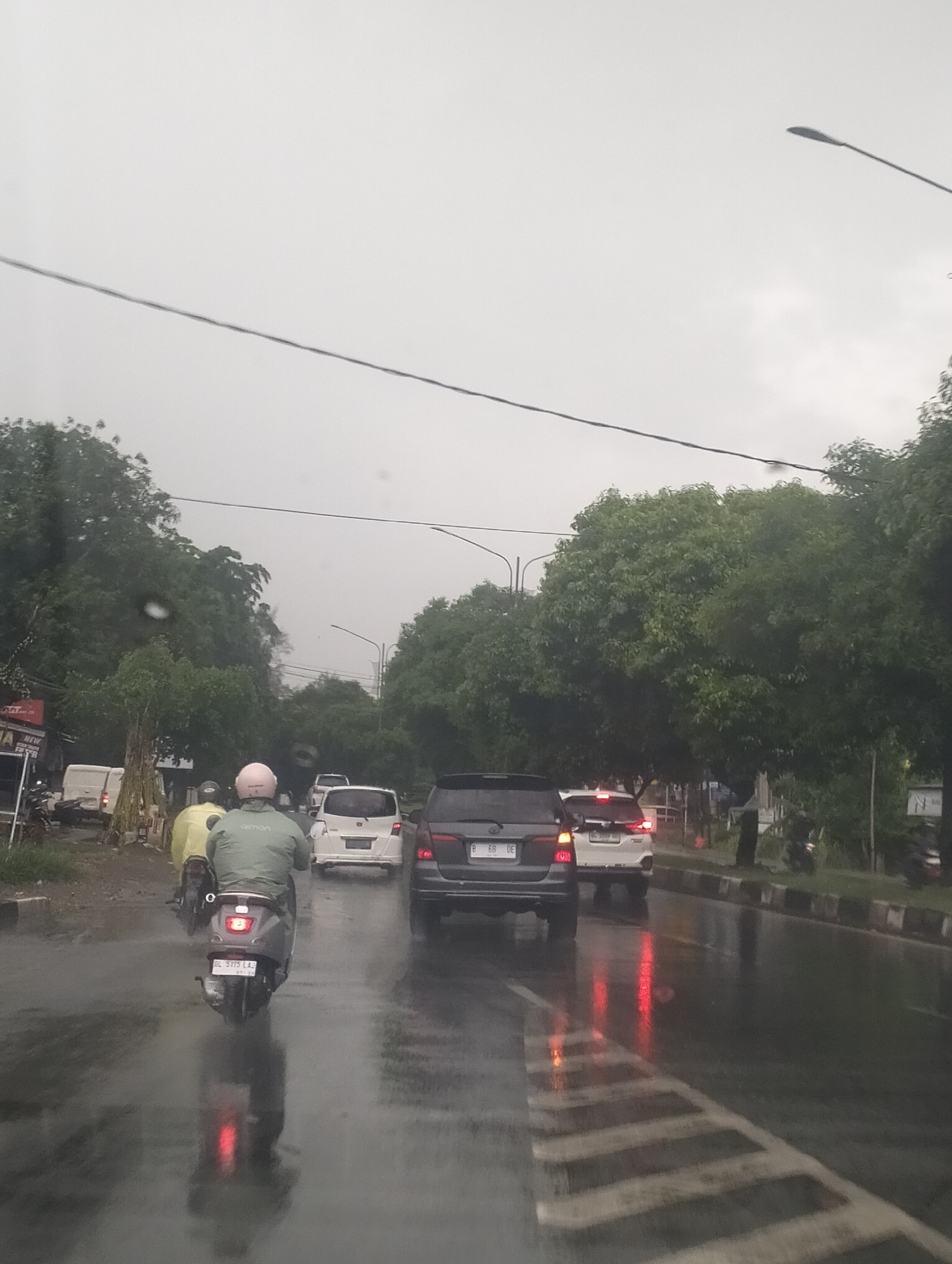 This is a vertical, eye-level shot of a rainy street scene in Meunasahmanyang, Indonesia. The weather is overcast, and the road is wet and reflective, with visible water droplets on the lens. A person on a scooter, wearing a light green jacket and a pink helmet, is in the foreground, facing away from the camera and traveling on the wet road. The scooter has a license plate that reads "BL 5775 LAJ".
Several cars are also on the road. Behind the scooter is a white car, and further ahead, a black SUV and another white car. The reflections of brake lights are visible on the wet surface of the road. The scene is lined with lush green trees on both sides of the road, with a few power lines running overhead. There are also streetlights visible in the background.
On the left side of the road, there are buildings and a white van. Some text is visible on a red sign to the left, partially obscured but appearing to say "NEW". The overall mood is damp and hazy due to the rain. The time of day appears to be late afternoon or early evening, given the dim, overcast sky.