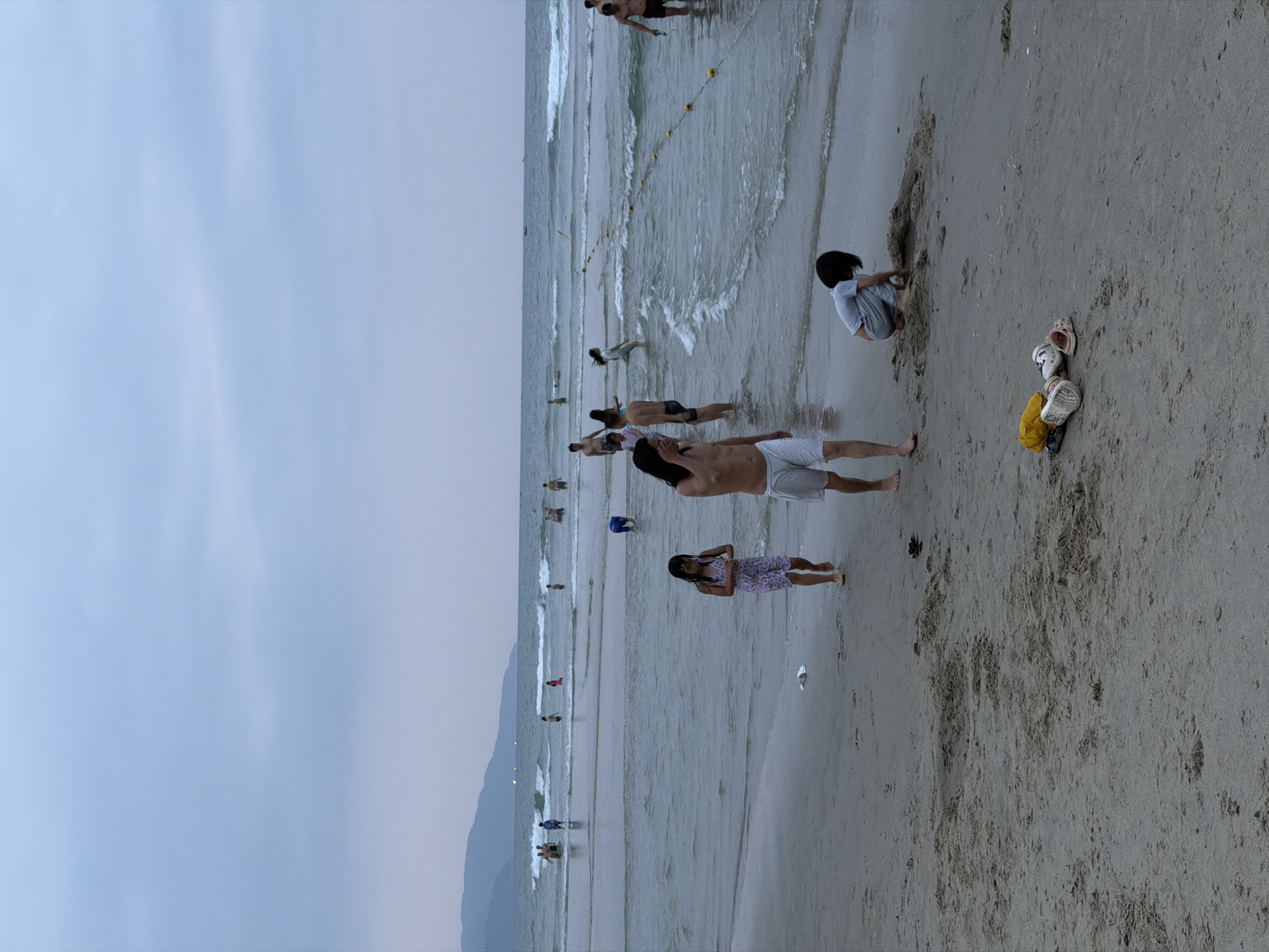 The image depicts a beach scene in Đà Nẵng, Vietnam. The sky is overcast. Several people are present, both in the water and on the sandy beach. In the water, some individuals are swimming or standing. On the beach, one person is digging in the sand, while another stands nearby. A set of shoes and a yellow bag are sitting in the sand. Further in the distance, a mountain range is visible, fading into the sky. The water has waves breaking near the shore. A safety rope with yellow floats stretches along the water.