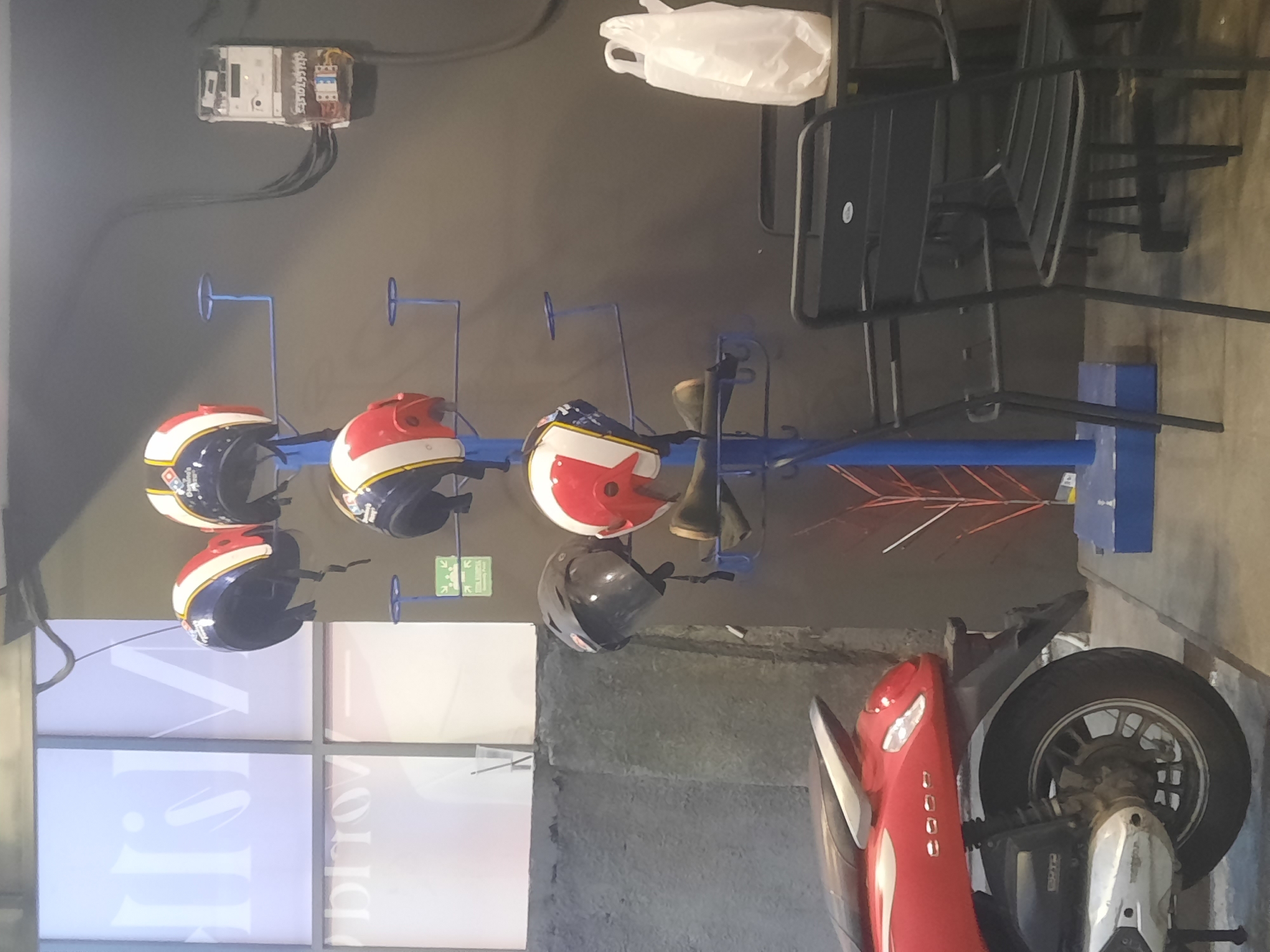 The image shows an indoor scene, likely a cafe or a waiting area, with a motorcycle parked in the foreground on the left. A blue helmet rack stands in the middle of the frame, holding several helmets. To the right, there are tables and chairs, with a white plastic bag on one of the tables. A gray wall serves as the backdrop, with a window and some electrical equipment visible. The lighting suggests an artificial light source. The location is North Jakarta, Indonesia.