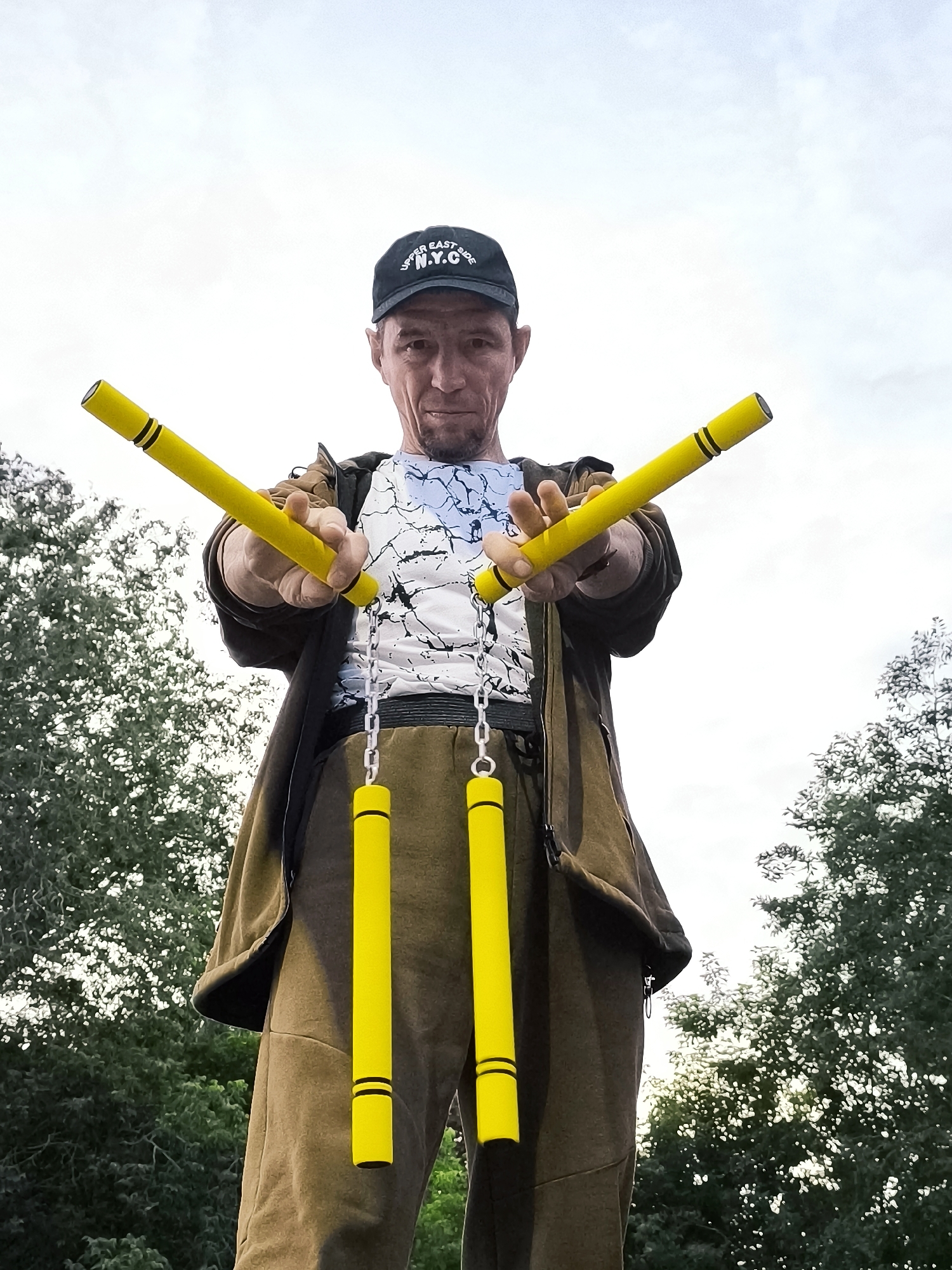 A man is shown from the chest up, holding a pair of yellow nunchaku. He is wearing a black baseball cap with "UPPER EAST SIDE N.Y.C." embroidered on it, a white t-shirt with a black abstract design, and a brown jacket. The man has short, grey hair and a goatee. He is standing outdoors with trees in the background and a cloudy sky above.